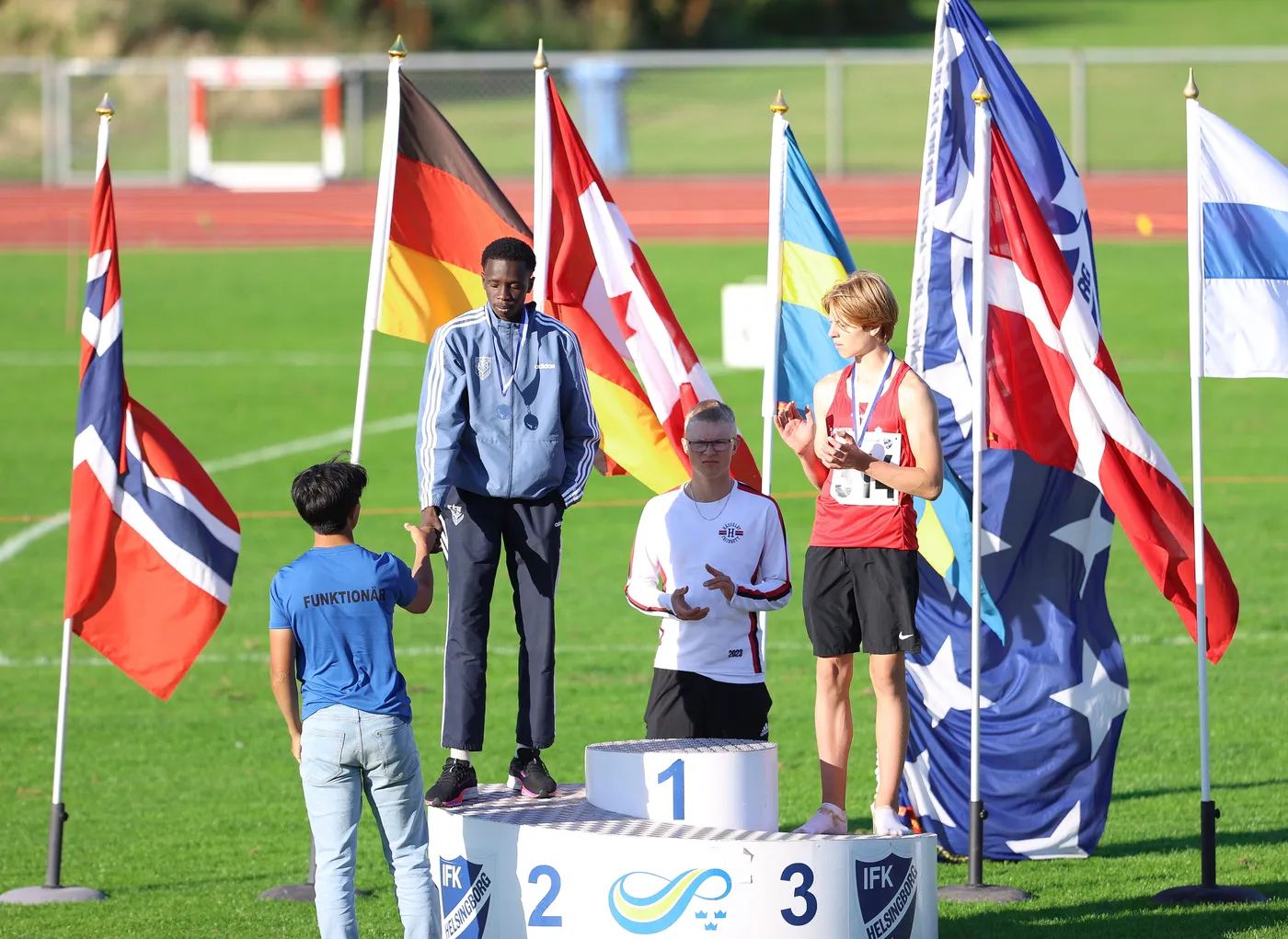 ثلاثة رياضيين يقفون على منصة التتويج مع الأعلام الوطنية المرفرفة من حولهم. الرياضي الذي يرتدي سترة زرقاء يحمل ميدالية في المركز الثاني، بينما الرياضي الذي يرتدي سترة حمراء يحصد المركز الأول. يتفاعل شخص آخر معهم وهو يرتدي قميصاً أزرق.