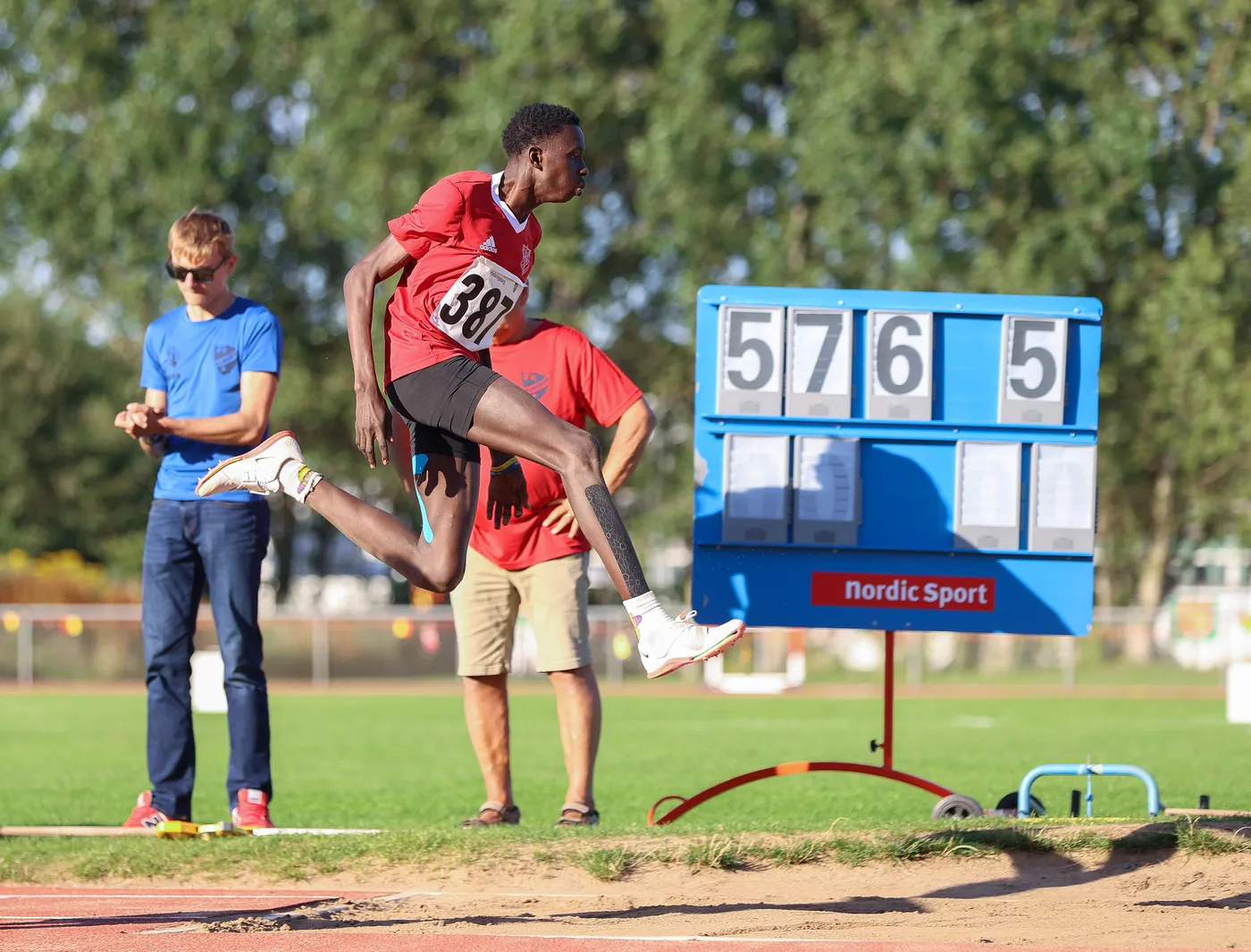 عداء يرتدي زيًا رياضيًا أحمر يحمل الرقم 381، يقوم بقفزة طويلة أثناء المنافسة. في الخلفية، يظهر اثنان من المشجعين، واحد منهم يرتدي نظارات شمسية، بالإضافة إلى لوحة تسجيل تعرض الأرقام 5765.