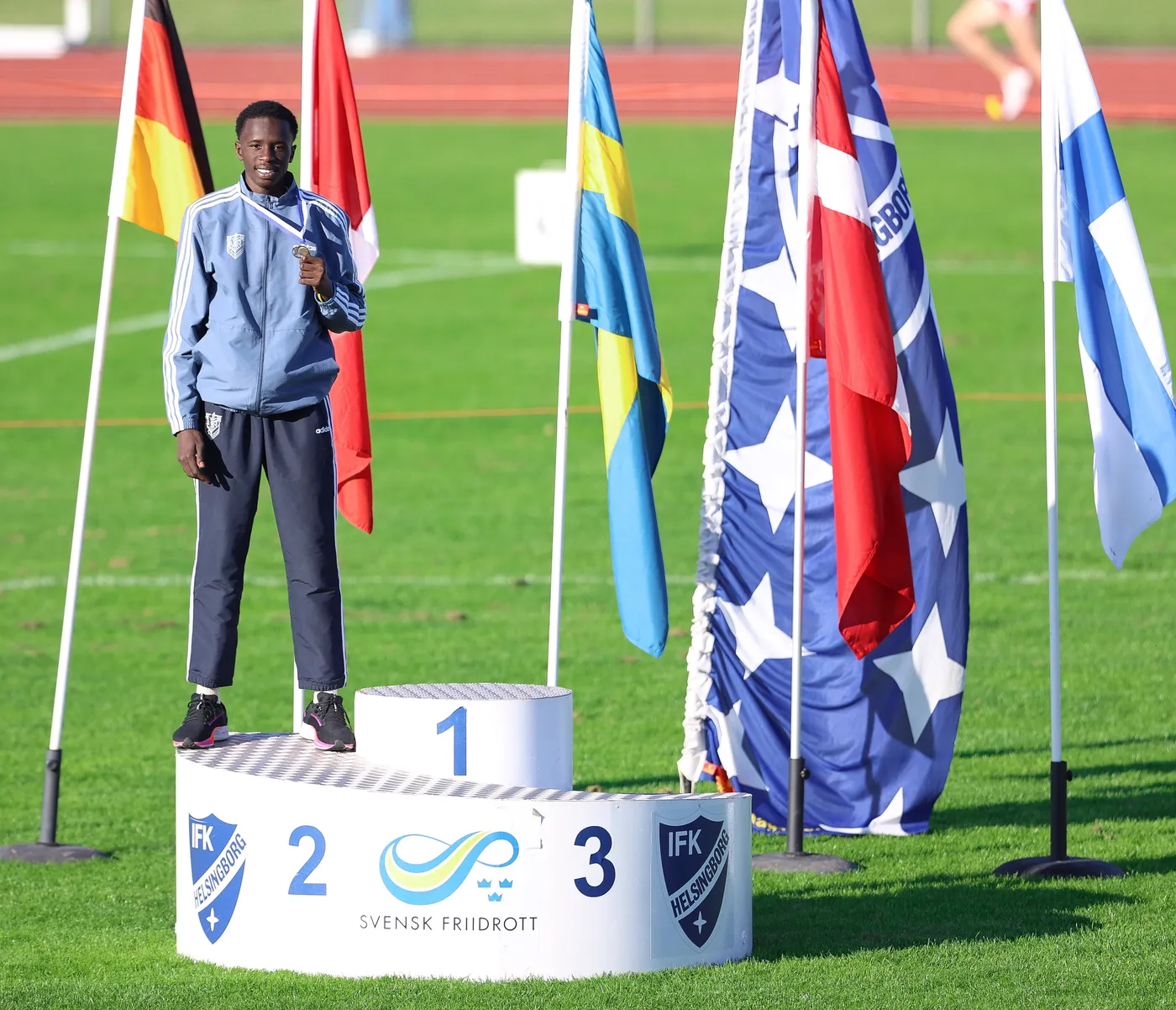 شاب يقف على المنصة للفائزين مع العلم السويدي ورايات دول أخرى من حوله. يحمل الميدالية في يده ويظهر سعيدًا.