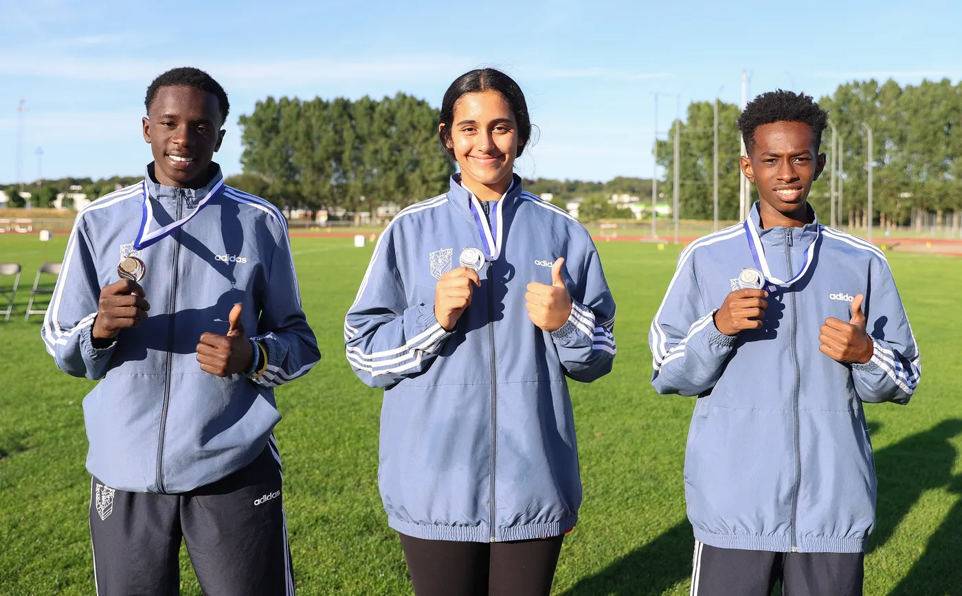 ثلاثة رياضيين يقفون في حقل رياضي، يرتدون جاكيتات رياضية زرقاء. يحمل أحدهم ميدالية برونزية بينما تظهر ميدالية فضية لدى الأخرى. جميعهم مبتسمون ويشيرون بإبهامهم إلى الأعلى إشارة للنجاح.