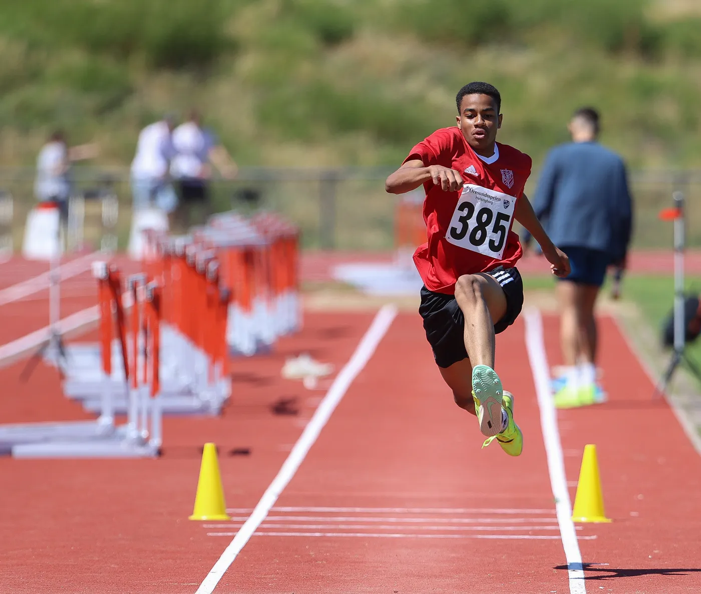 رياضي يرتدي زيًا أحمر مع رقم 385، في وضعية القفز في مسابقة ألعاب القوى. يحيط به ميدان رياضي مع مجموعة من الحواجز وأعلام ملونة، وأشخاص آخرين في الخلفية.