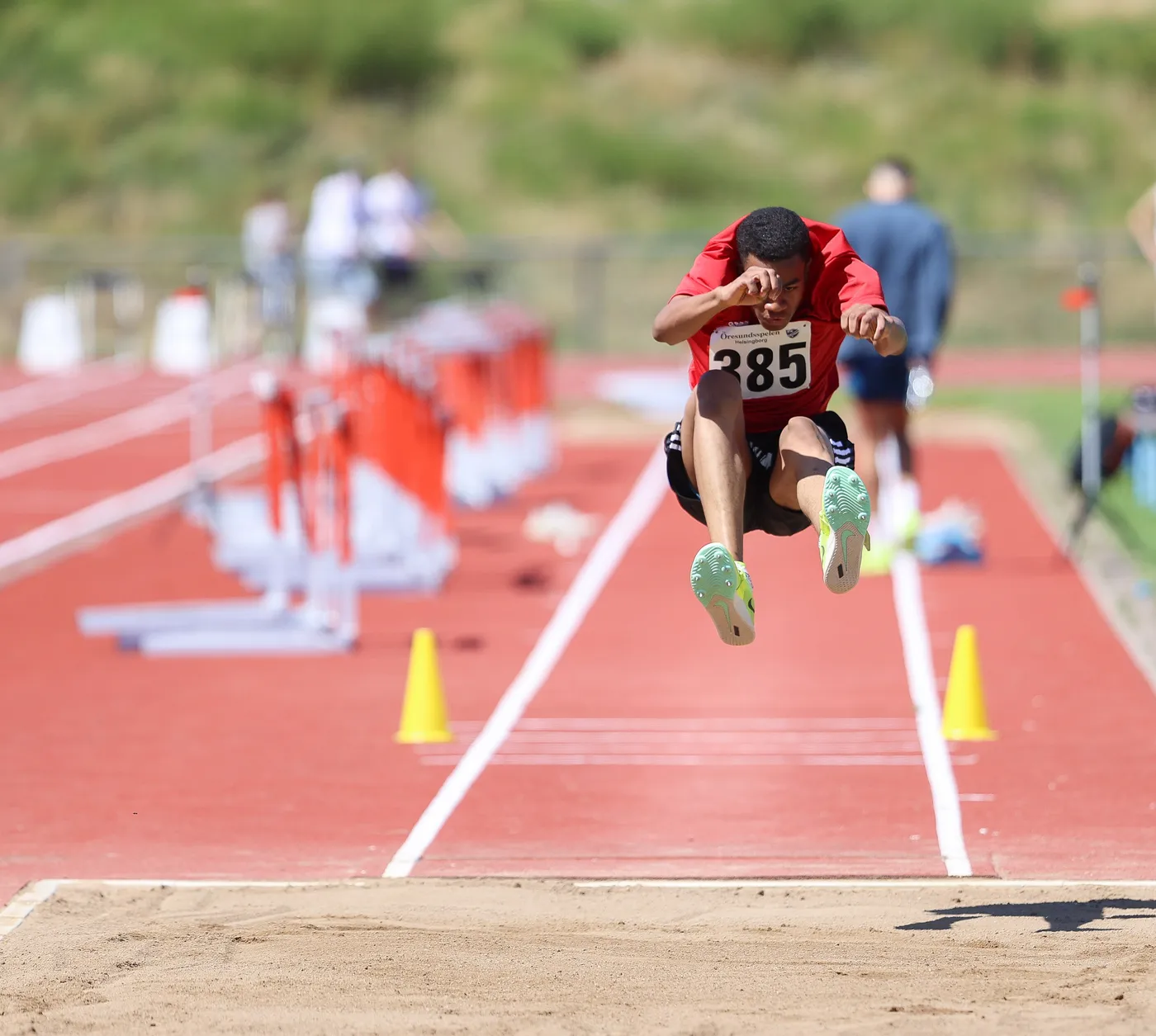 لاعب عداء يقفز لأعلى خلال منافسة، مرتديًا قميصًا أحمر ورقم 385، فوق حلبة رياضية ذات لون أحمر. في الخلفية، توجد حواجز رياضية وأشخاص مشغولون بالأنشطة الرياضية.
