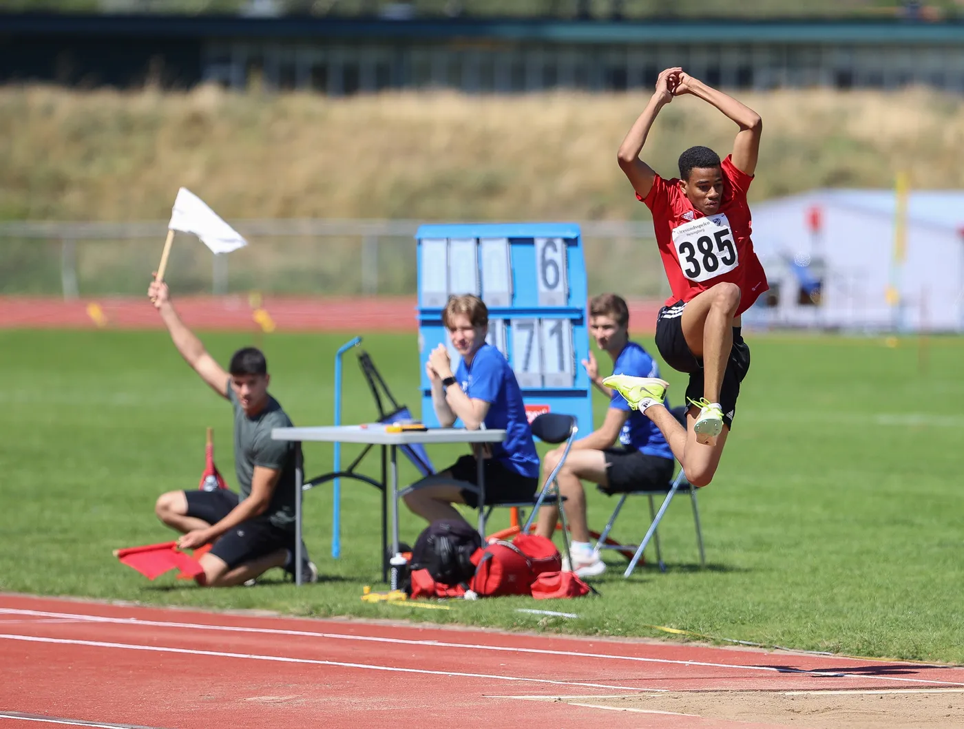 يشارك شاب في مسابقة الوثب الطويل، وهو يرتدي قميصًا أحمر يحمل الرقم 385. في الخلفية، يظهر حكام ومسؤولون عن المسابقة يجلسون على طاولة، بينما يشير أحدهم بعلم أبيض.