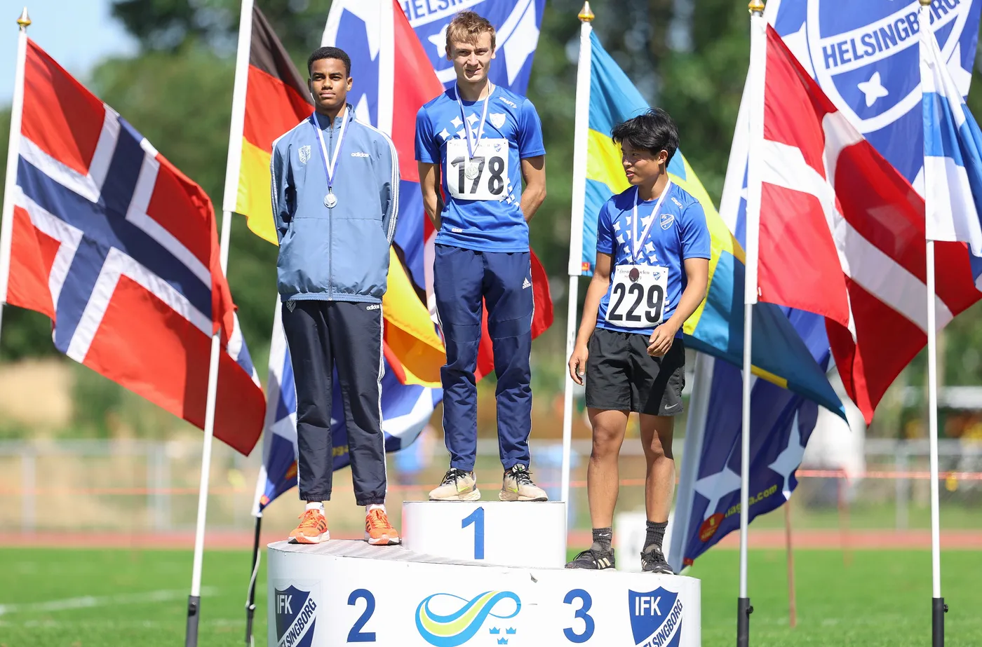 ثلاثة متسابقين يقفون على منصة التتويج، حيث يحمل الفائز بالمركز الأول ميدالية ذهبية، بينما يحمل المتسابقان الآخران ميداليتين فضية وبرونزية. تحيط بهم أعلام متعددة الدول، مما يعكس أجواء المنافسة الرياضية.