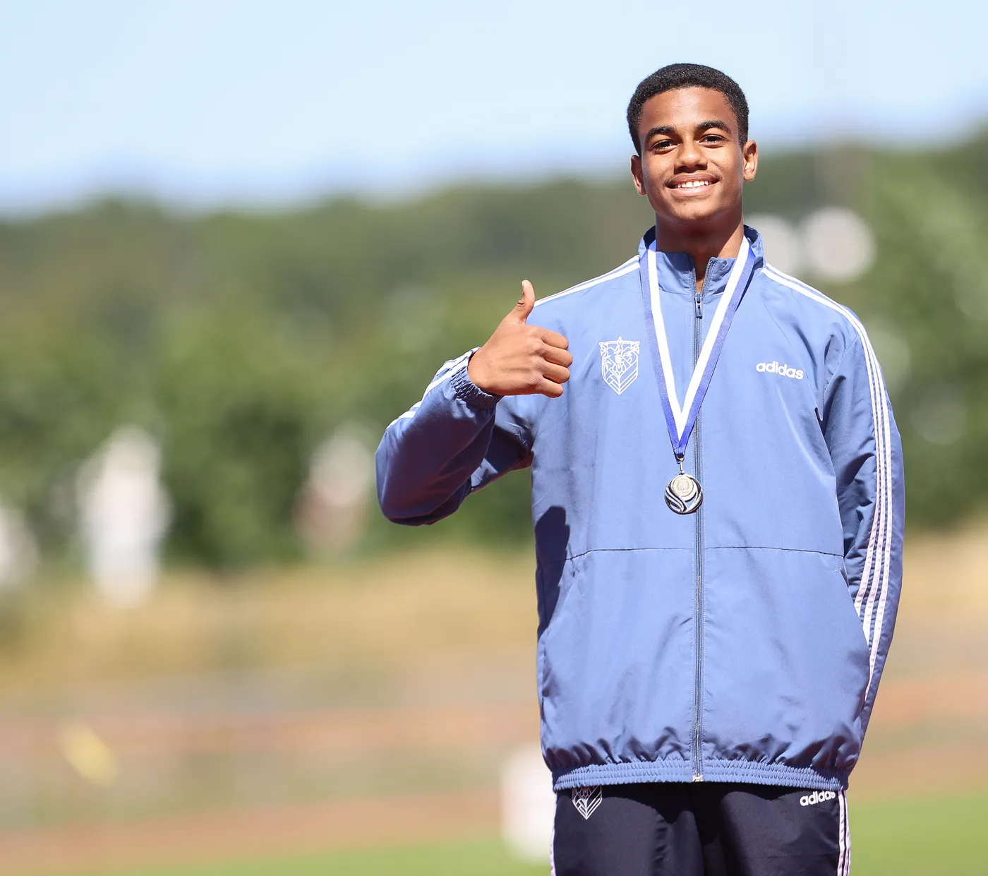 شاب مبتسم يرتدي معطف رياضي أزرق وعليه ميدالية فضية، يشير بإبهامه لأعلى كدلالة على الفخر. الخلفية تظهر أرضية رياضية مع بروز للون الأخضر.