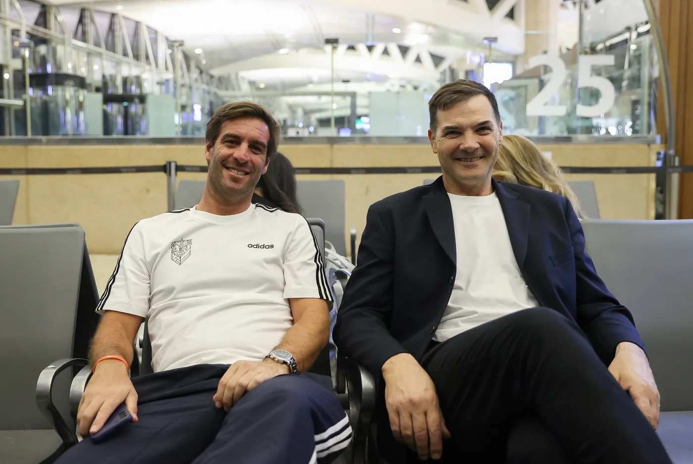 رجلان يجلسان معًا مبتسمين في مطار حديث. يرتدي أحدهما قميص رياضي أبيض مع علامات تجارية، بينما يرتدي الآخر بدلة داكنة.