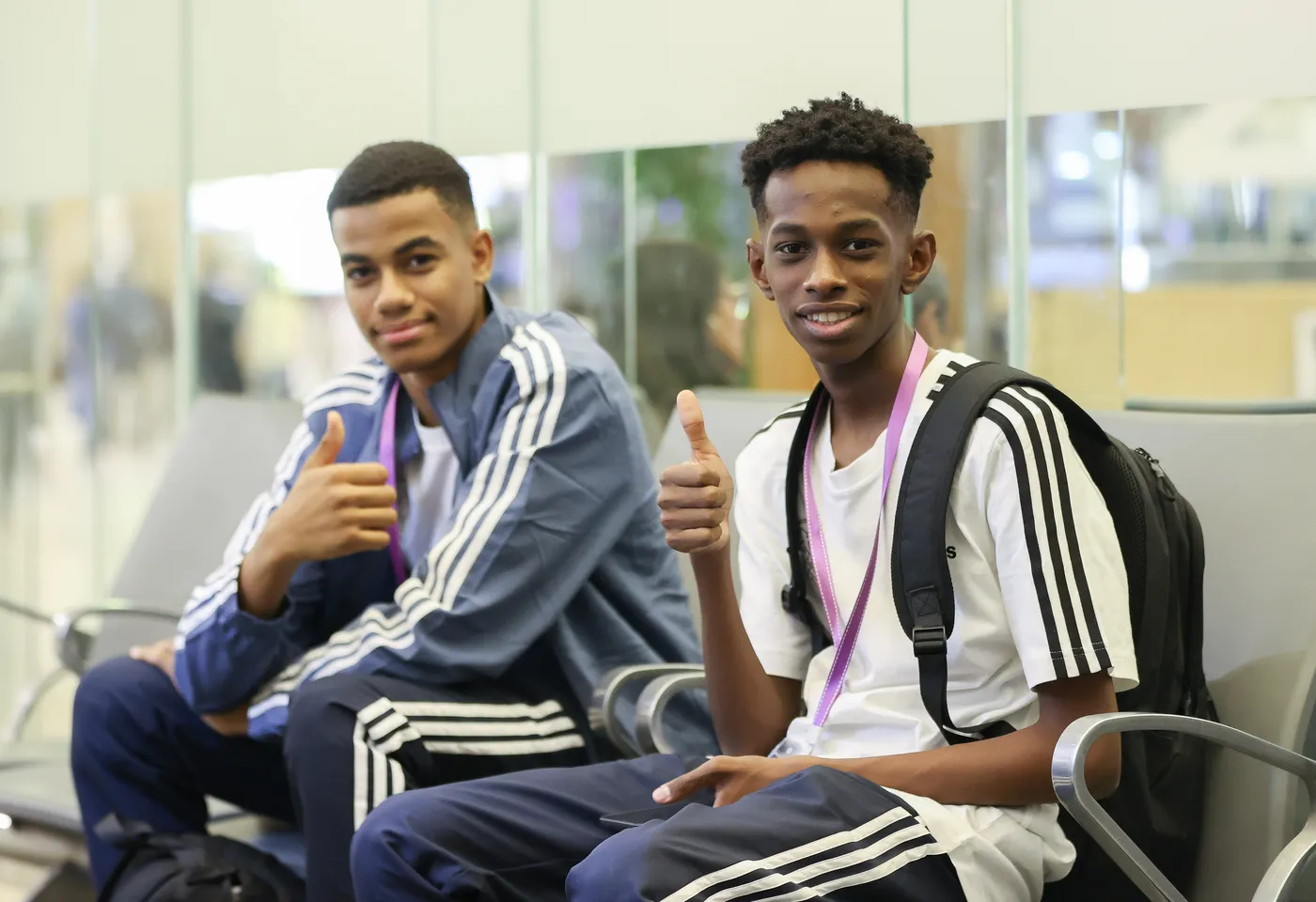 شابان يجلسان في منطقة انتظار. كليهما يرتدي ملابس رياضية ذات خطوط سوداء وبيضاء، ويبتسمان بينما يرفع كل منهما إبهامه للأعلى كإشارة على الرضا.