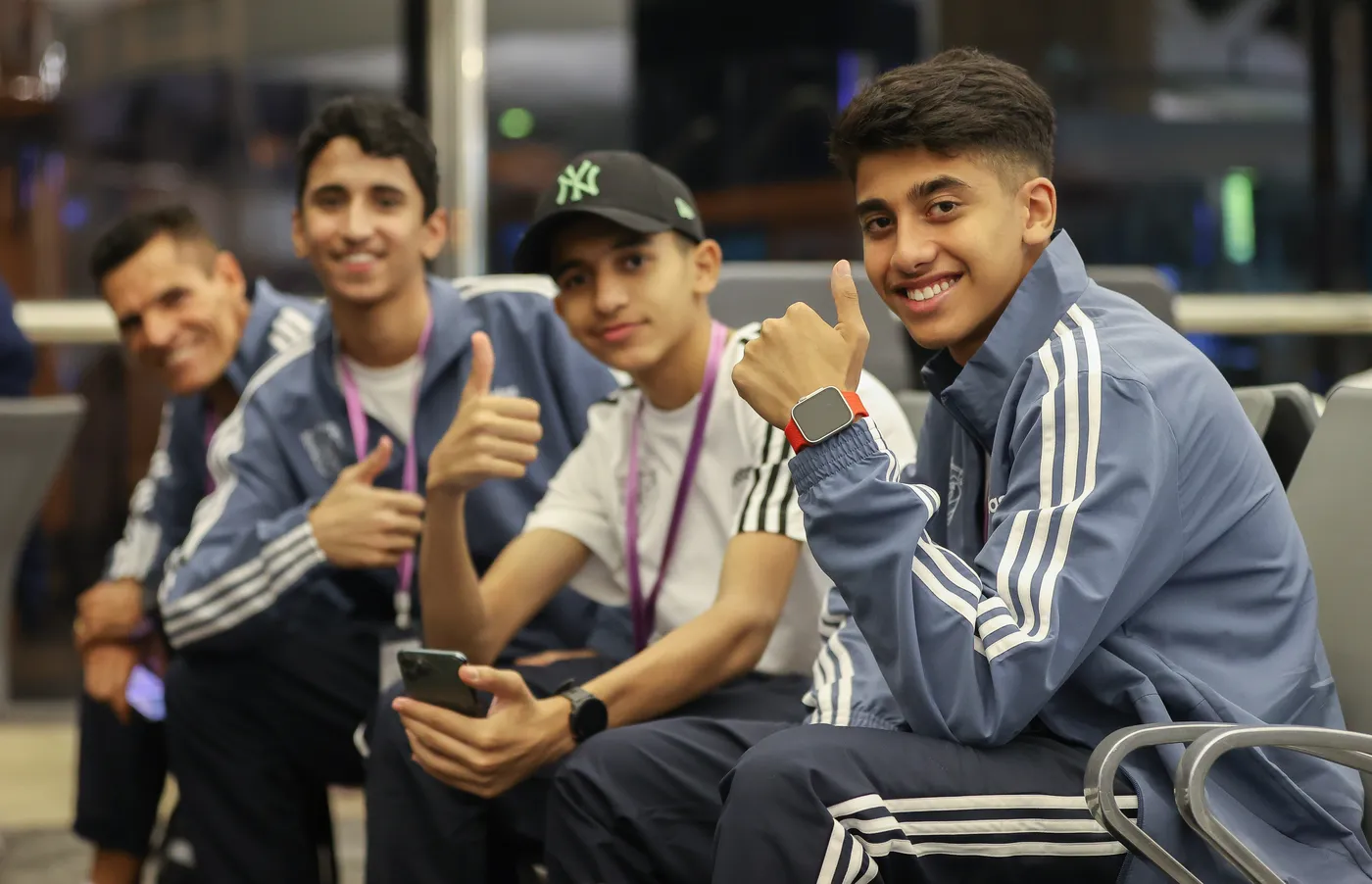 مجموعة من الشبان يجلسون معًا في مكان مريح، يبتسمون ويعرضون إشارات الإبهام إيجابية. يرتدون ملابس رياضية زرقاء، ويظهر في الخلفية شخص بالغ يبتسم أيضًا.