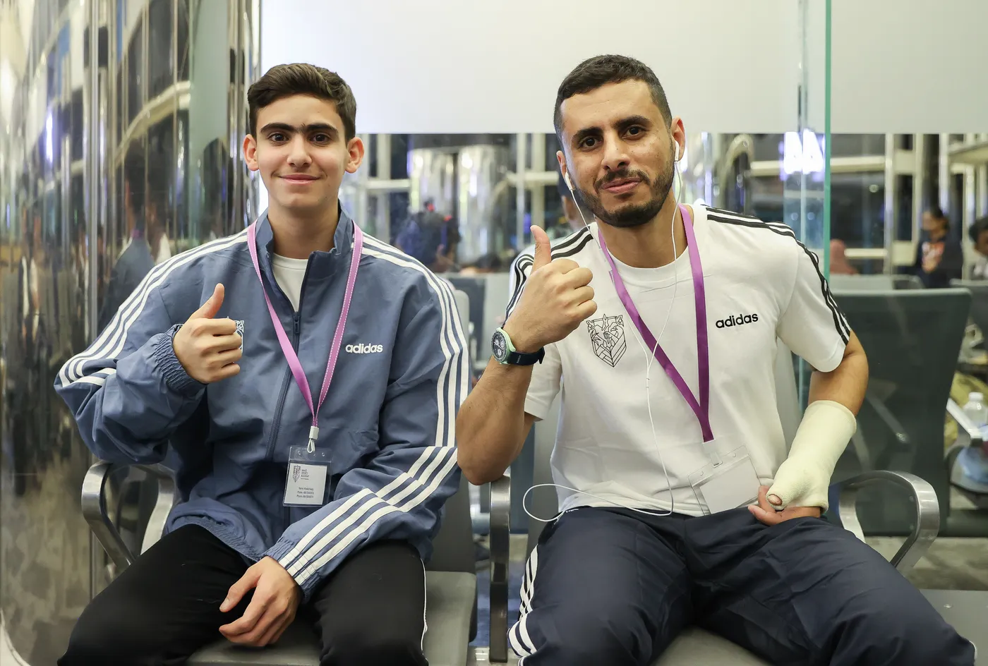 شابان يجلسان بجوار بعضهما البعض في مكان مغلق، يحمل كل منهما إصبع الإبهام للأعلى. يرتدي الأول سترة زرقاء، بينما يرتدي الثاني قميصًا أبيض مزينًا بشعار. كلاهما يرتدي شارات تعريفية، ويبدو أنهما في وضع مريح.