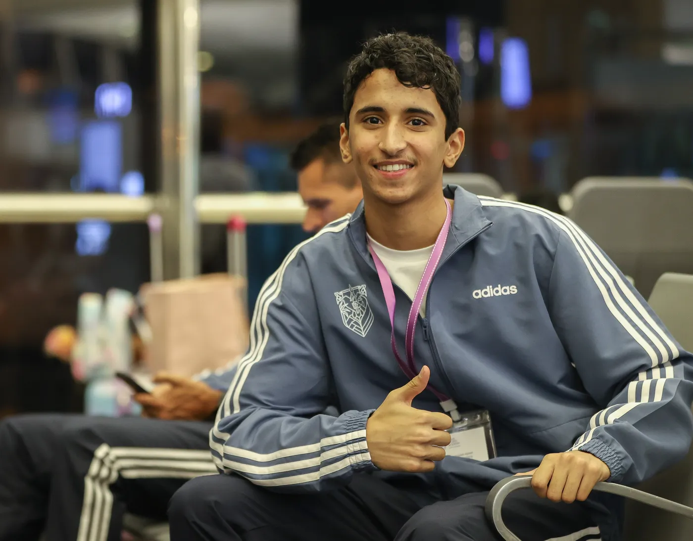 شاب مبتسم يجلس على كرسي، مرتديًا سترة رياضية ذات لون أزرق ورمز علامة تجارية معروفة. تظهر يداه في وضع الإبهام المرفوع، مما يعكس شعورًا بالراحة والسعادة، بينما يجلس شخص آخر خلفه.