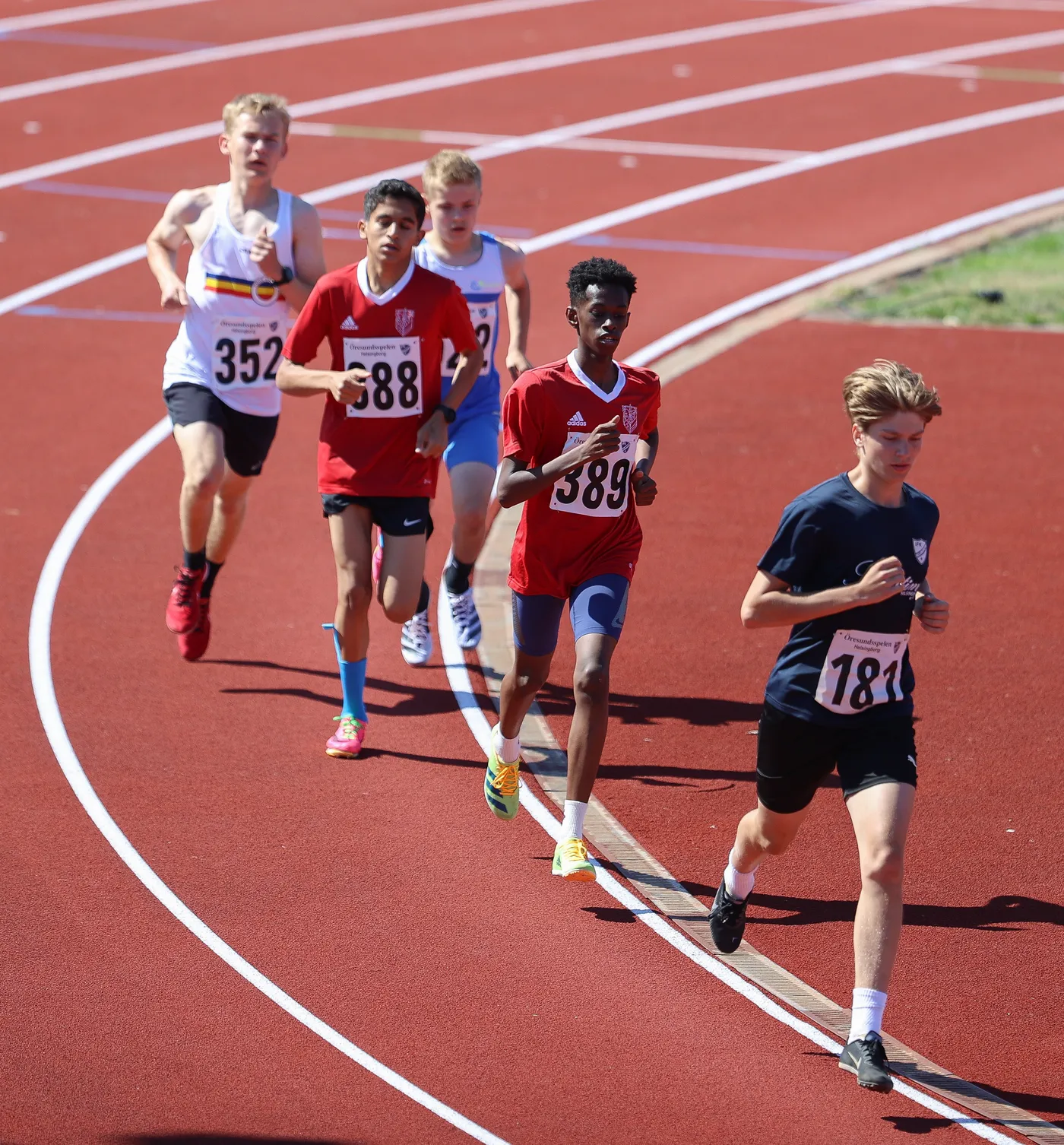 مجموعة من المتسابقين الشباب يتنافسون في سباق على مضمار ألعاب القوى. يرتدي بعضهم قمصان حمراء بينما الآخرون يرتدون ألواناً متنوعة، ويظهرون في مرحلة متقدمة من السباق.