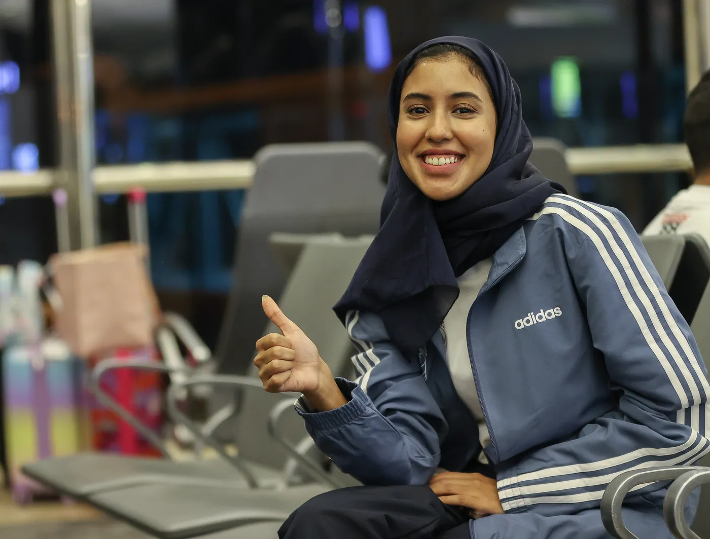 امرأة شابة ترتدي زي رياضي أزرق وتضع حجابًا، تجلس على مقعد في مكان مخصص للانتظار وتبتسم. تشير بإصبعها البارز إلى الأعلى، تعكس شعورًا بالسعادة والثقة.