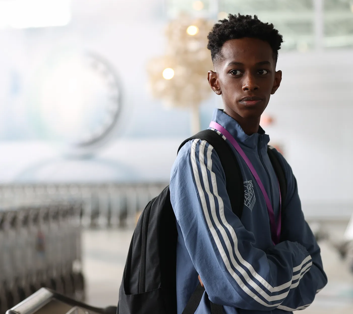 شاب ذو تسريحة شعر قصيرة ومجعد، يرتدي سترة زرقاء بحواف بيضاء وحقيبة ظهر، يقف في مكان مفعم بالضوء. يبدو جاداً ويركز على شيء ما خارج إطار الصورة.