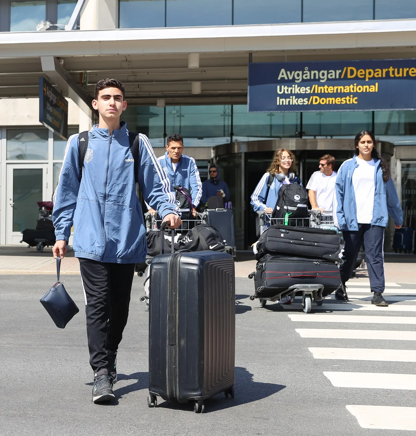 مجموعة من الشباب ترتدي ملابس رياضية زرقاء، يسيرون نحو مخرج مطار. يحمل أحدهم حقيبة سفر كبيرة بينما يحمل آخرون عربات لحقائبهم.