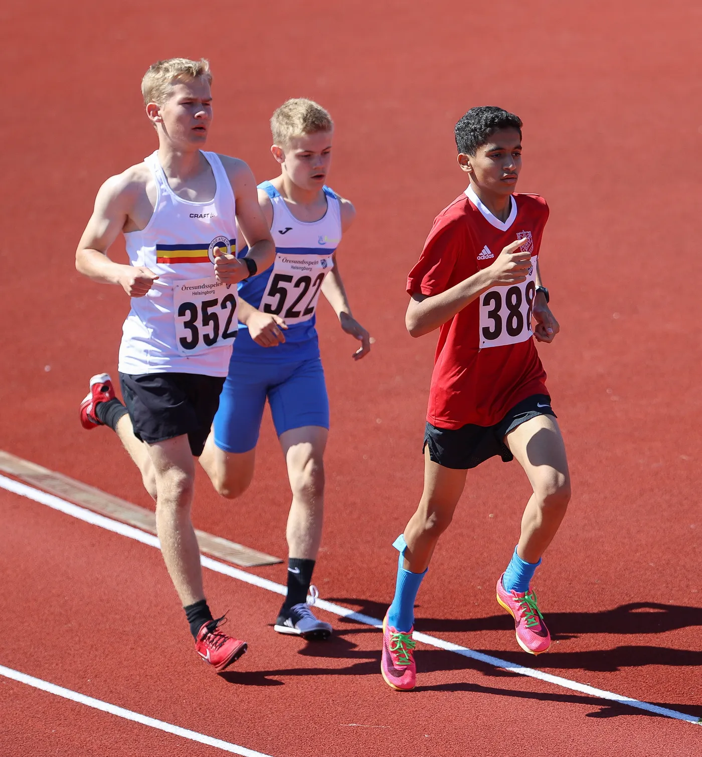 ثلاثة عدائين يتنافسون على مضمار رياضي أحمر. يرتدي العداؤون قمصاناً رياضية بألوان مختلفة، حيث يحمل كل منهم رقمًا على صدره.