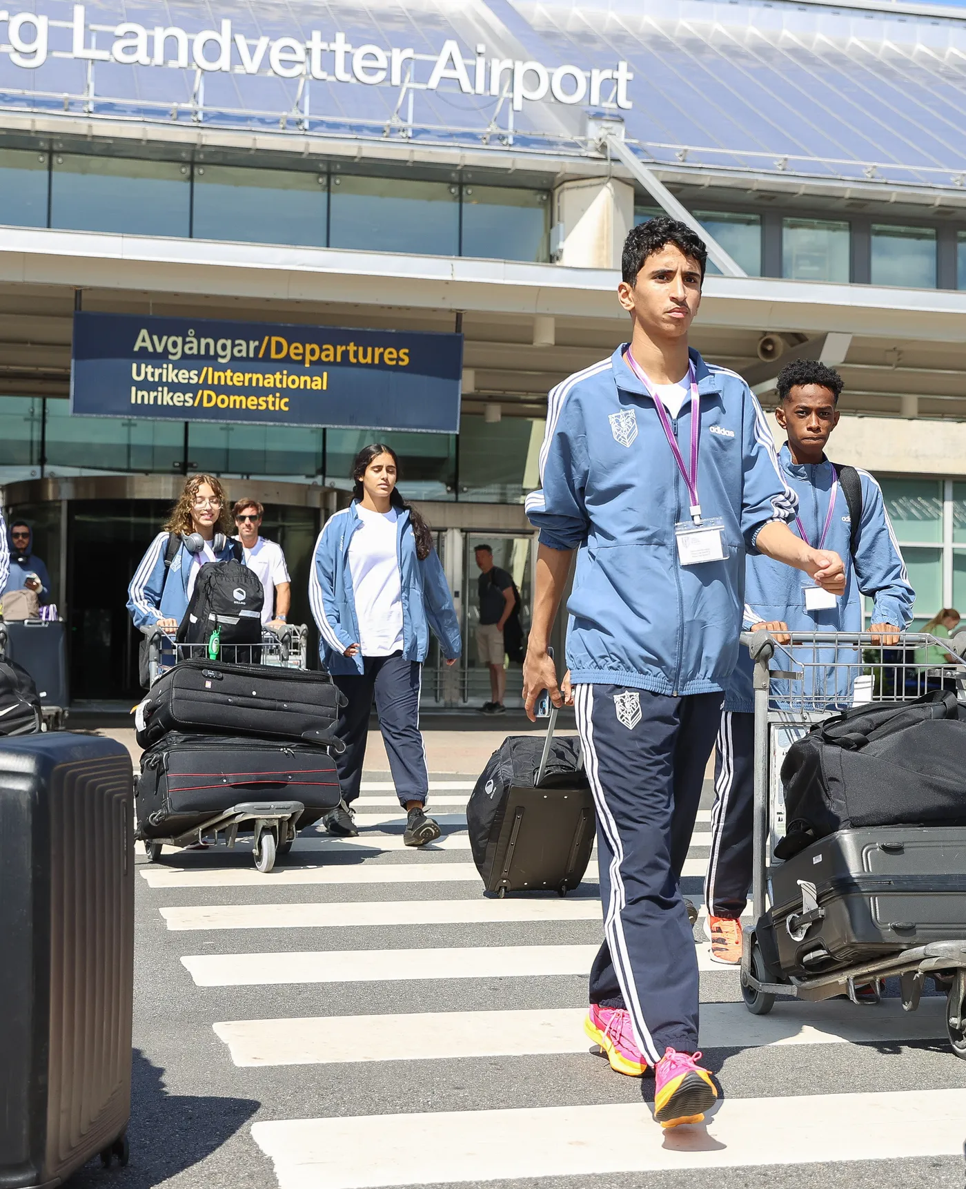 مجموعة من الشباب يرتدون ملابس رياضية وهم يسيرون نحو المطار، مع عربات تحمل الحقائب الثقيلة. تُظهر الإضاءة الساطعة يومًا مشمسًا في مطار لاندفيتي.