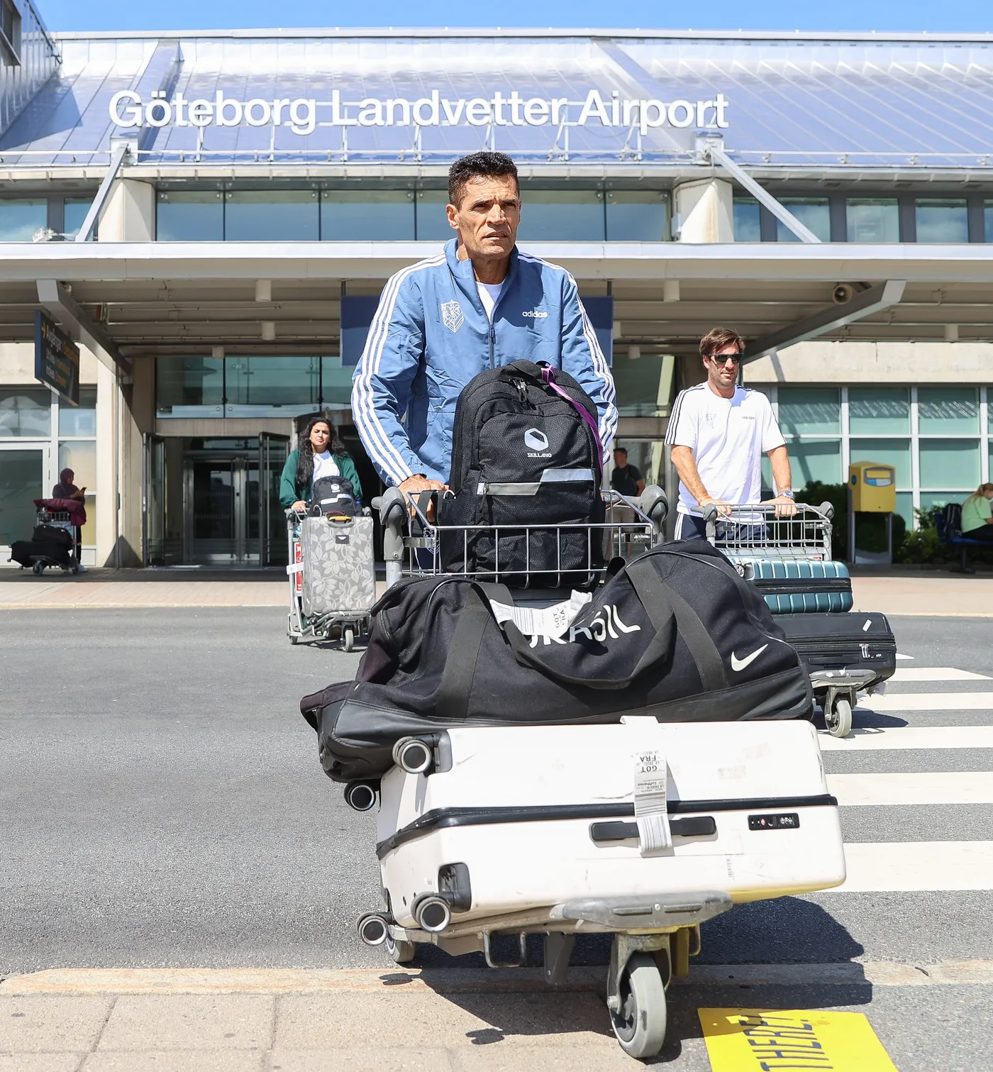 رجل يدفع عربة أمتعة في مطار غوتنبيرغ لاندفتر، يحمل حقيبة ظهر سوداء وأمتعة كبيرة، بينما يظهر مسافرون آخرون خلفه. يُرى لافتة تشير إلى المطار في الخلفية.