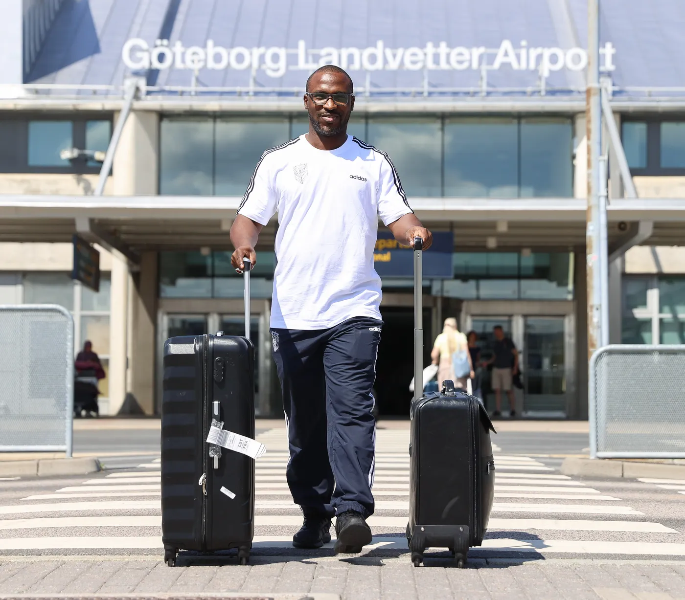 رجل يحمل حقيبتين على عجلات ويسير نحو مدخل مطار غوتنبيرغ لاندفيتر. يرتدي قميصًا أبيض وبنطالًا رياضيًا، ويبدو مبتسمًا تحت أشعة الشمس.