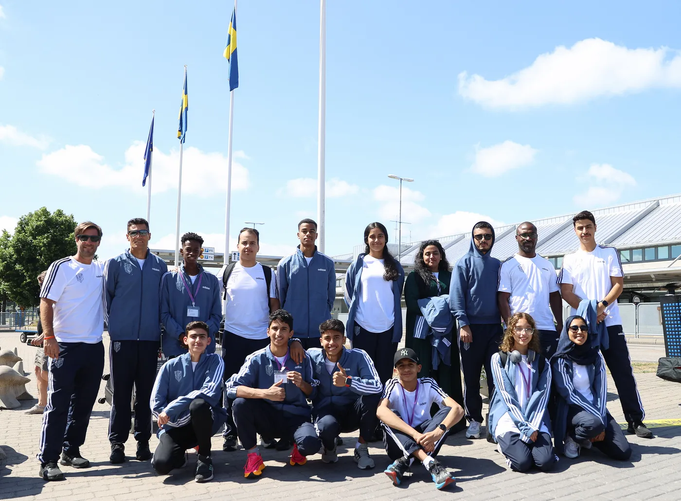 مجموعة من الشباب والبالغين يقفون معًا في منطقة خارجية تحت السماء الزرقاء، حيث ترفرف الأعلام في الخلفية. يرتدي بعضهم ملابس رياضية تتضمن سترات زرقاء وتيشيرتات بيضاء، بينما يظهر آخرون بملابس عادية. المجموعتان تتكونان من فتيان وفتيات، بعضهم مبتسم ويظهر علامات السعادة.