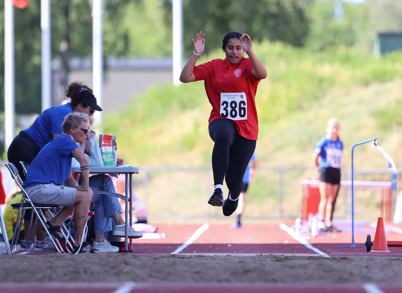 طفلة ترتدي قميصًا أحمر يحمل الرقم 386، أثناء قفزها في مسابقة القفز الطويل. في الخلفية، يظهر عدد من الأشخاص يجلسون على طاولة، وممارسون آخرون في الملعب.