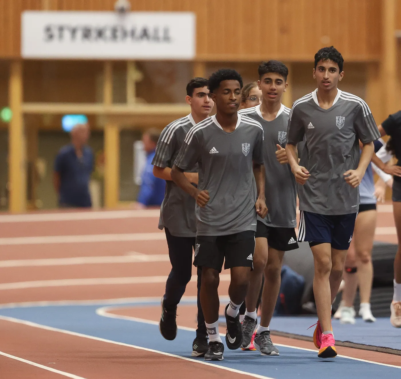 مجموعة من الشبان يرتدون قمصاناً رياضية باللون الرمادي يتمرنون في مضمار داخلي. يظهرون في حالة نشاط وجاهزية للتمرين، ويبدو عليهم التركيز والجدية.