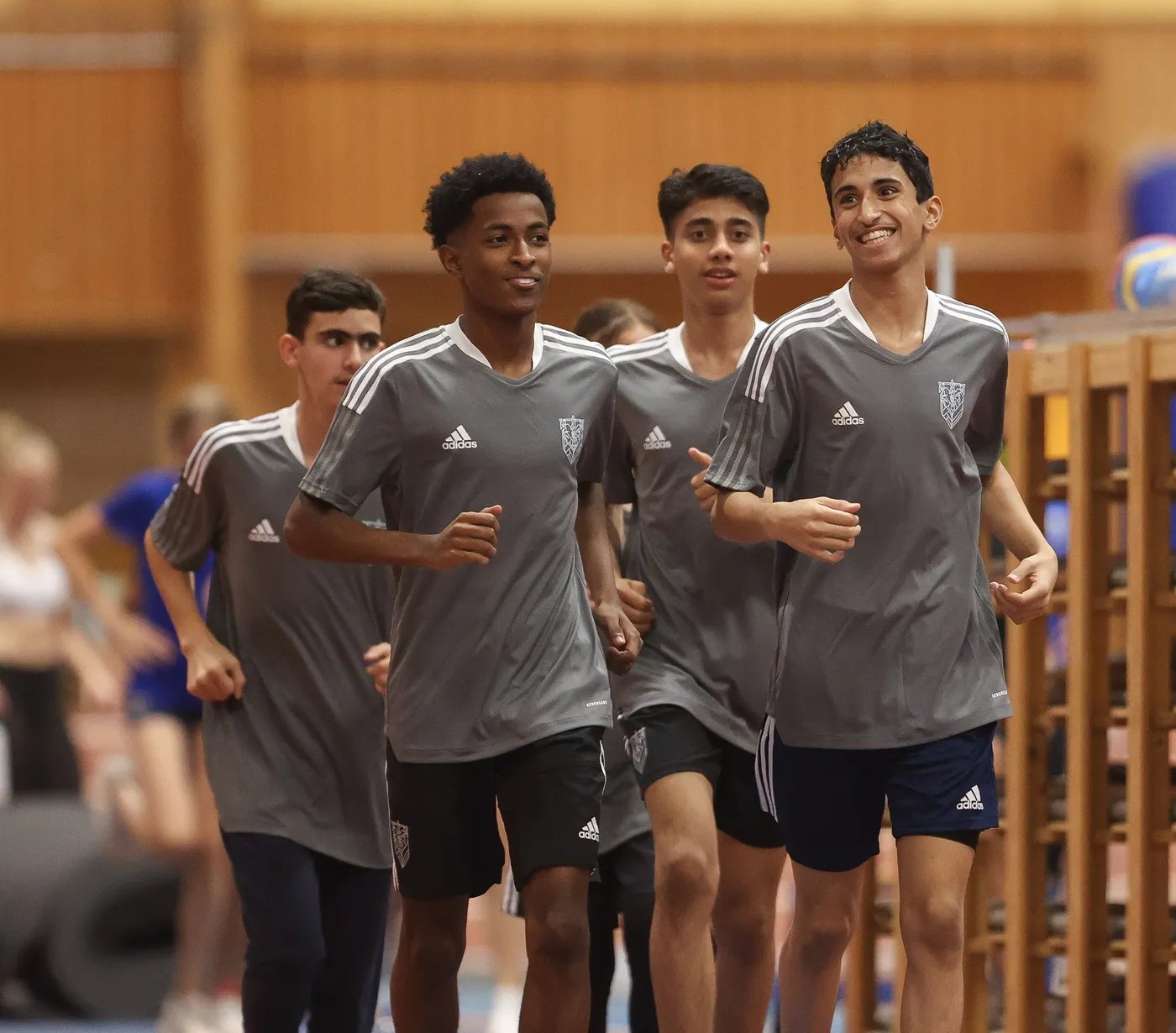 مجموعة من الشباب يمارسون الجري في صالة رياضية، جميعهم يرتدون قمصان رياضية رمادية اللون بحواف بيضاء. يظهر البعض منهم مبتسمًا، في حين يتابع آخرون الجري بجدية.