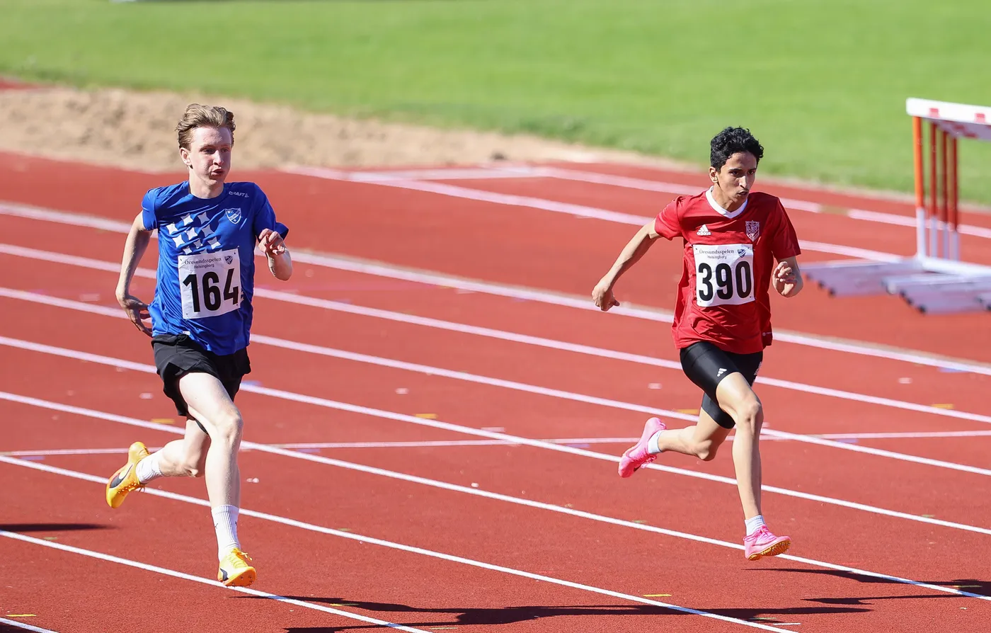 شابان يتسابقان في مضمار ألعاب القوى، الأول يرتدي أكمامًا قصيرة زرقاء مع رقم ١٦٤، والثاني بقمصان حمراء مع رقم ٣٩٠. الخلفية خضراء مع مضمار رياضي بلون أحمر.