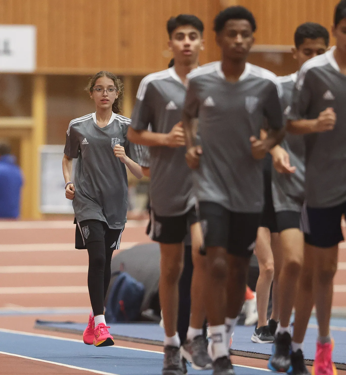 مجموعة من الرياضيين الشباب يرتدون قمصاناً رمادية قصيرة أثناء الجري في حلبة داخلية. الفتاة الوحيدة في المقدمة ترتدي حذاءً وردياً وتبدو مركّزة.
