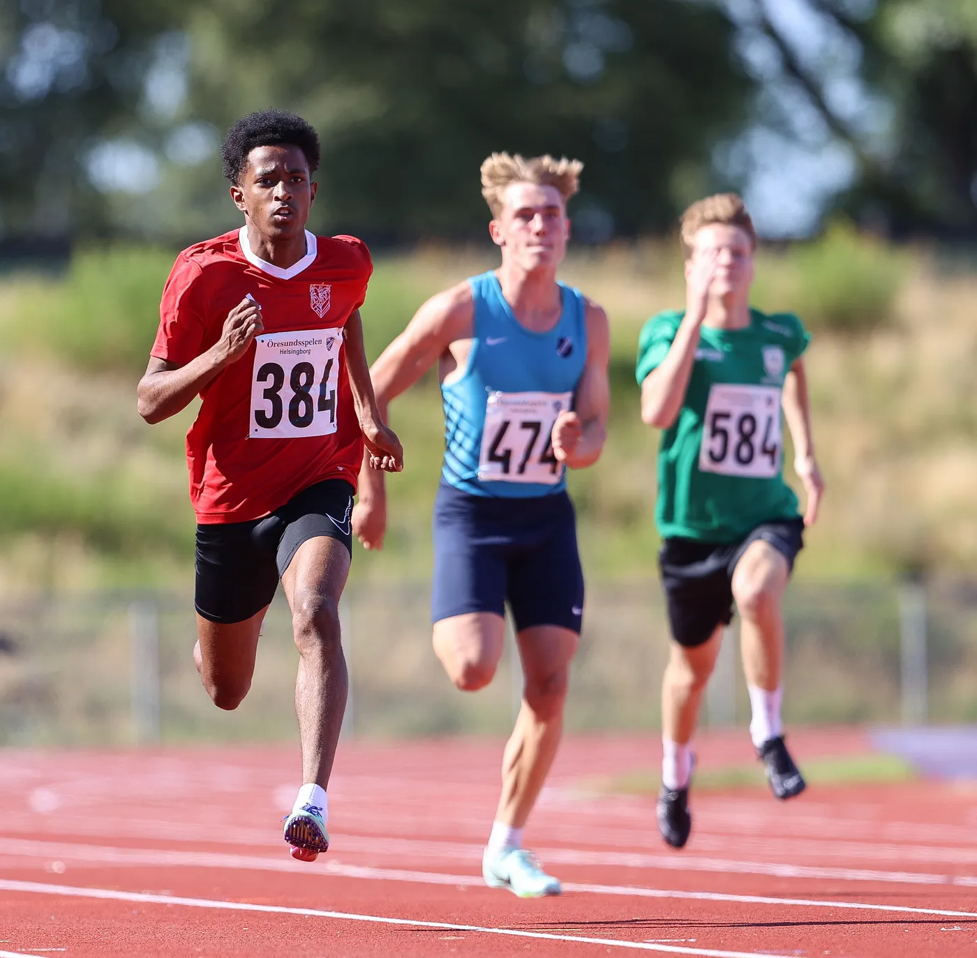 عداء يرتدي قميصاً أحمر ورقم 384 يسبق عدائين آخرين خلال مسابقة جري على مضمار رياضي. تظهر خلفه مجموعة من المتسابقين كل منهم يرتدي زي مختلف وأرقام مختلفة.