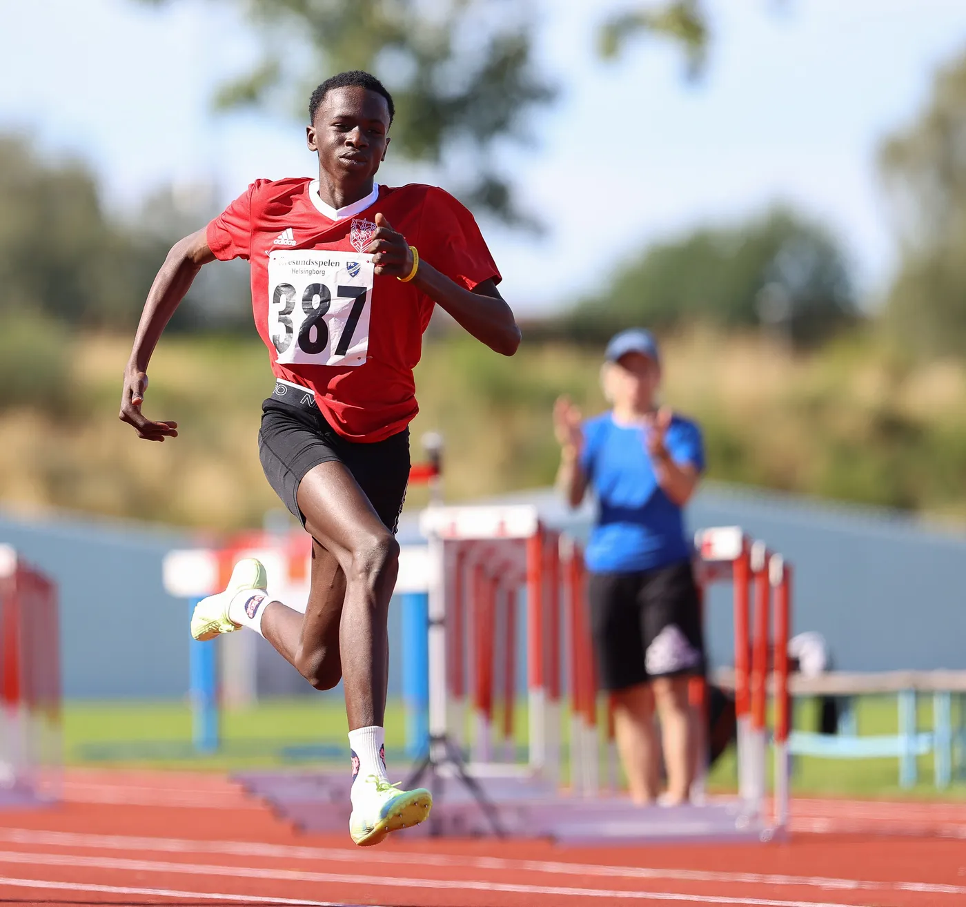 عداء يرتدي قميصًا أحمر برقم 387، يتسابق على حلبة ألعاب القوى، مع تعبير عن التركيز والحماس. في الخلفية، يظهر شخص يرتدي قميصًا أزرق يصفق له.