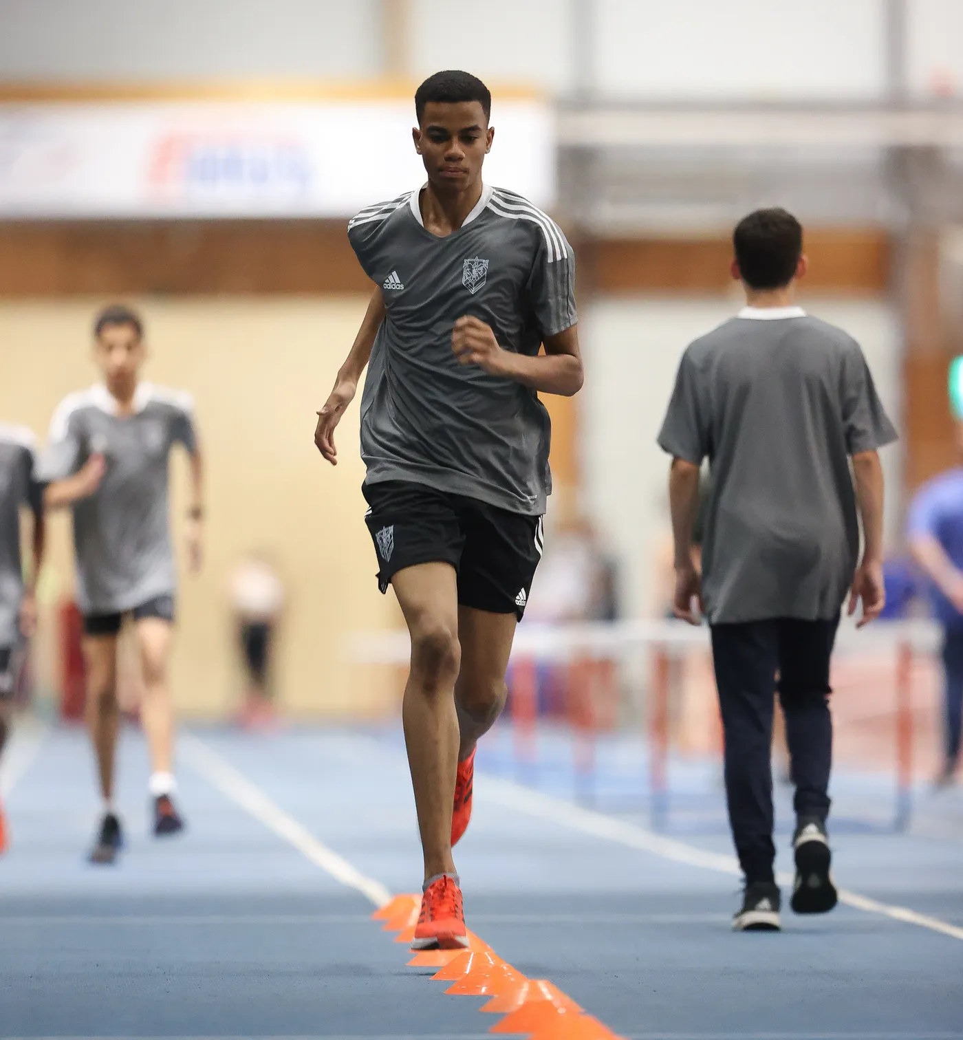 شخص شاب يركض في مضمار رياضي داخل قاعة، يرتدي زي رياضي رمادي مع شورت أسود وأحذية رياضية برتقالية. يوجد في الخلف مجموعة من الشبان أيضاً، بعضهم يمشي بينما يواصل الآخرون التمرين.