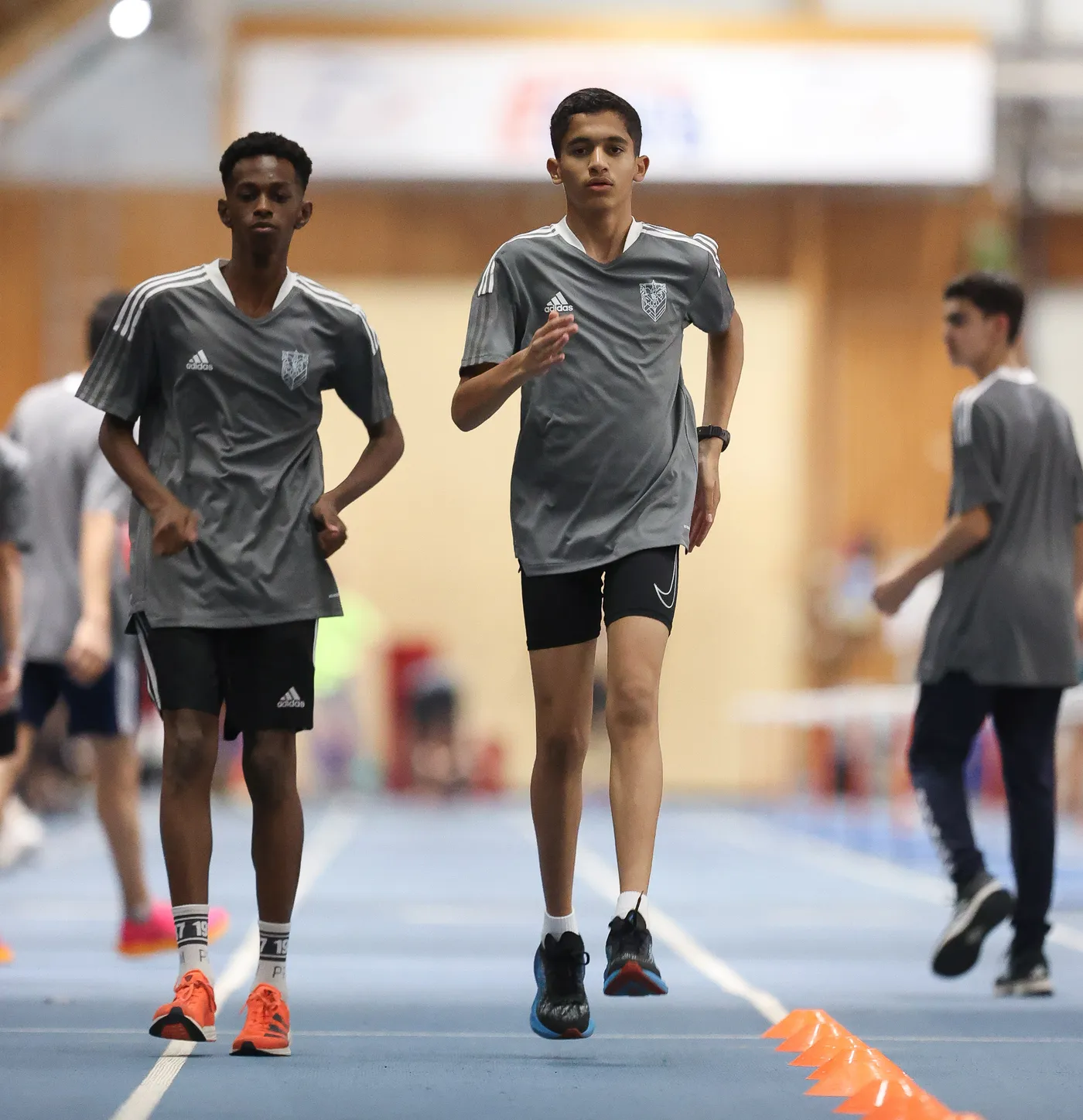 يقوم شابان بممارسة الجري في قاعة رياضية، حيث يرتديان قمصانًا رمادية وشورتات رياضية. يظهر في الخلف مجموعة من الشباب وهم يمارسون الرياضة أيضًا.