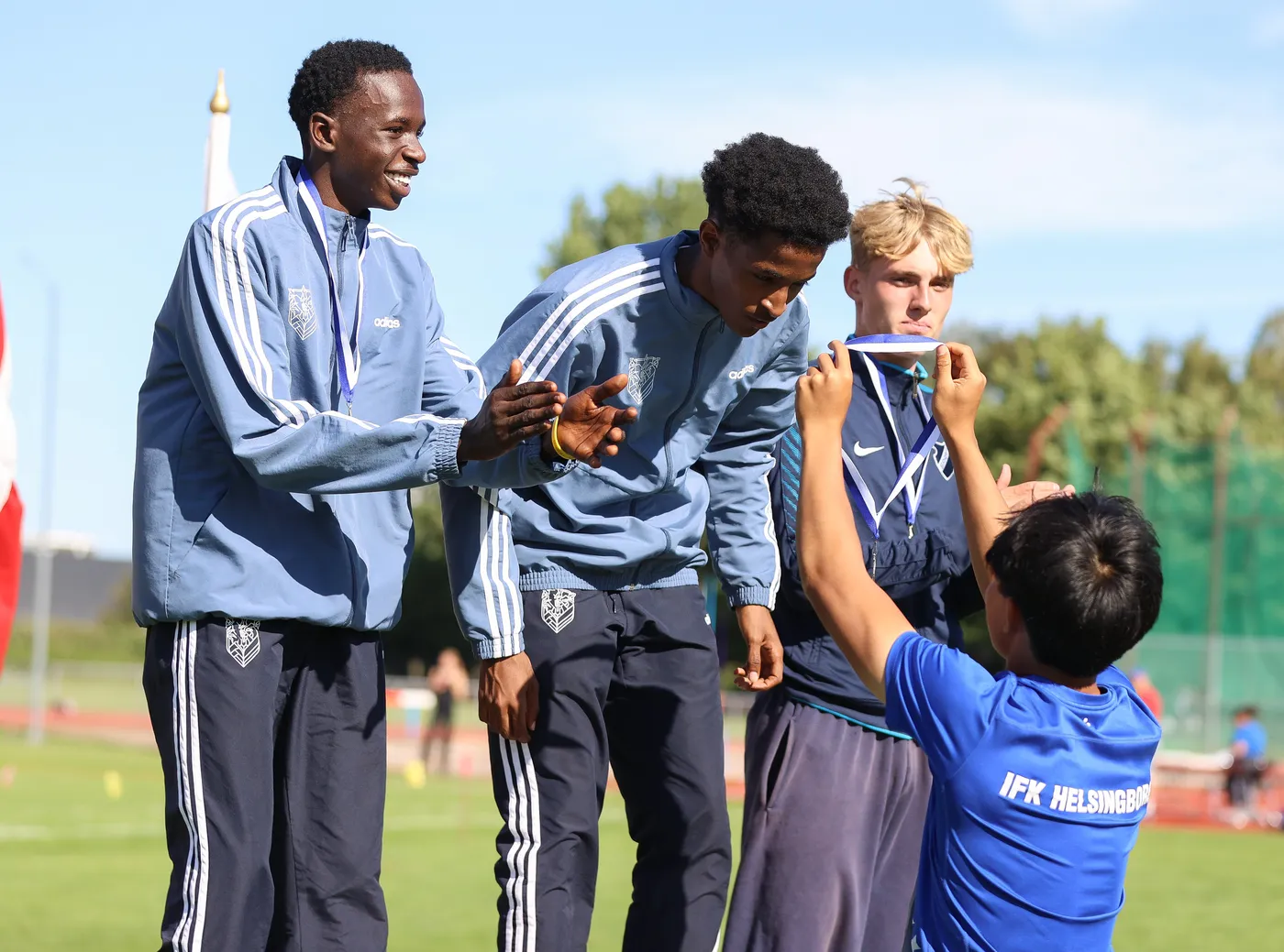 أربعة شبان يرتدون ملابس رياضية، حيث يحتفل أحدهم بتسلم ميدالية من شاب آخر يجلس أمامه. يظهر في الخلفية ملعب رياضي شاسع مع أشجار في الأفق. الجو يبدو مشمسًا.