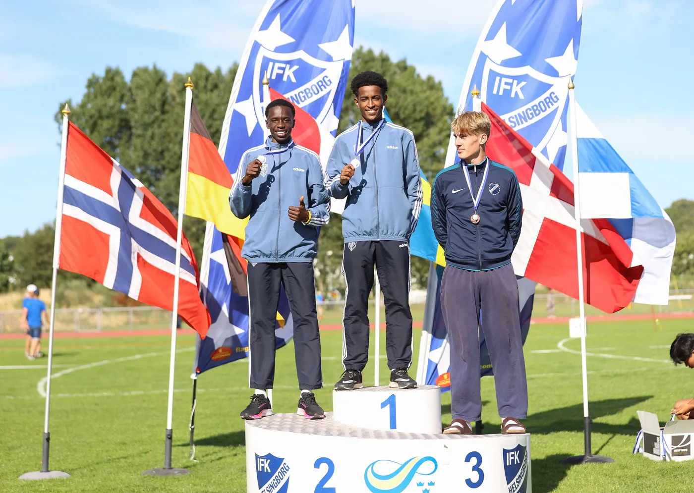 تجمّع ثلاثة رياضيين على منصة التكريم حيث يُظهر الأول فوز الميدالية الذهبية، والوسطى يحمل الميدالية الفضية، بينما الأخير يمثل الميدالية البرونزية. تُحيط بهم أعلام متعددة من دول مختلفة تشير إلى خلفية دولية للحدث.