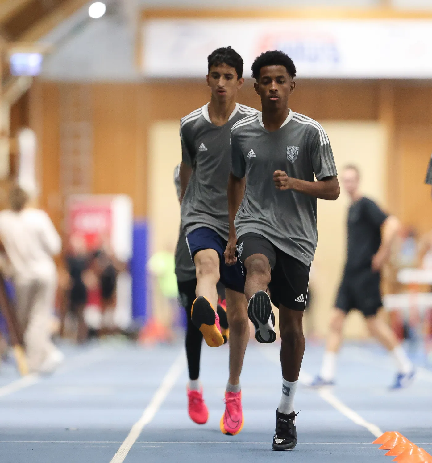 شبان يجريان على مضمار داخلي، يرتدون قمصاناً رياضية رمادية مع شعار، حيث يرفع أحدهم ركبته أثناء الركض. خلفهم، يوجد أشخاص آخرون يجتمعون في الصالة الرياضية.