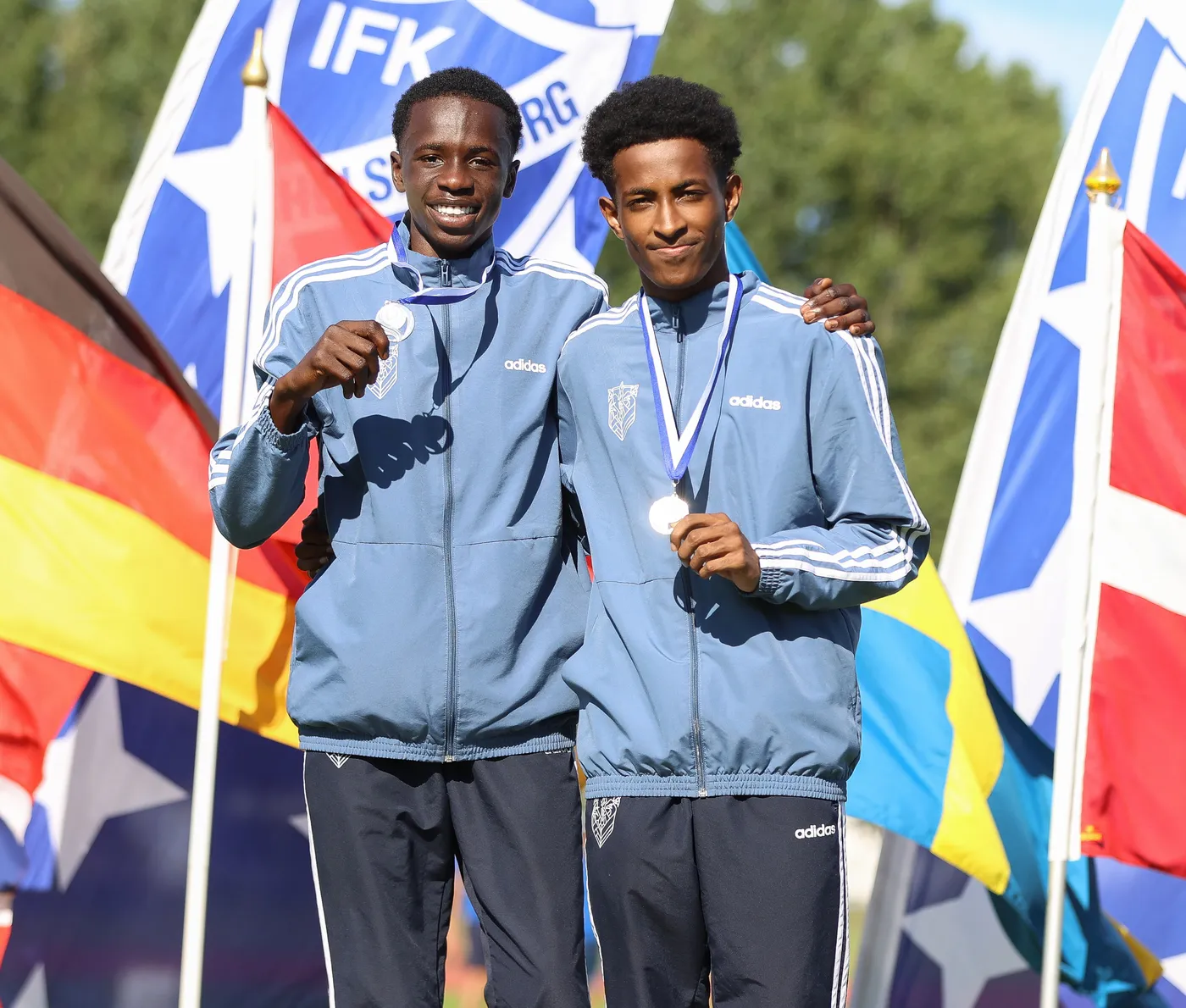 شابان يرتديان زيًا رياضيًا من شركة أديداس، يبتسمان معًا وهما يحملان ميداليات. خلفهما توجد أعلام متعددة لدول مختلفة، تعكس أجواء احتفالية.