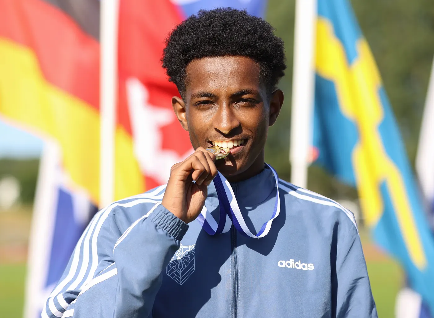 شاب يبتسم ويحمل ميدالية ذهبية في فمه، يرتدي سترة رياضية زرقاء تحمل شعار أديداس. خلفه مجموعة من الأعلام الملونة.
