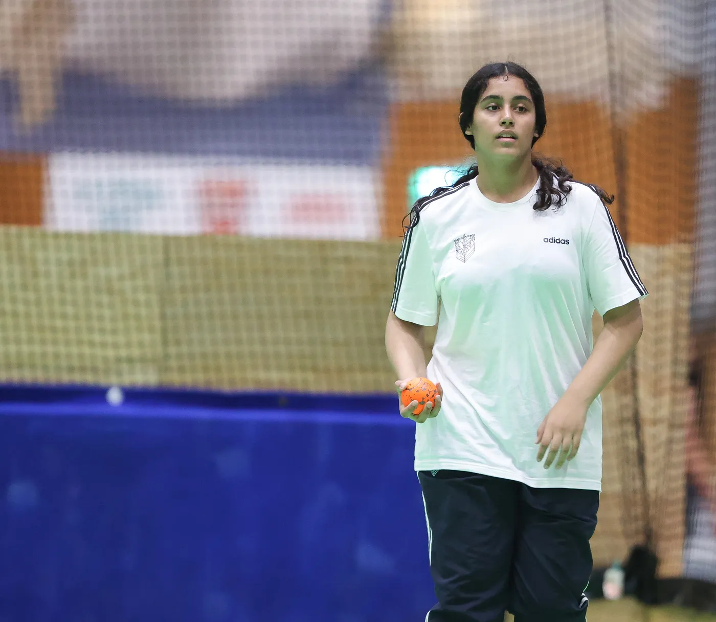 شابّة ترتدي ملابس رياضية بيضاء وسوداء، تحمل كرة برتقالية في يدها، وتظهر في صالة رياضية. خلفها شبكة رياضية، مما يوحي بأنها تستعد للمشاركة في لعبة رياضية.