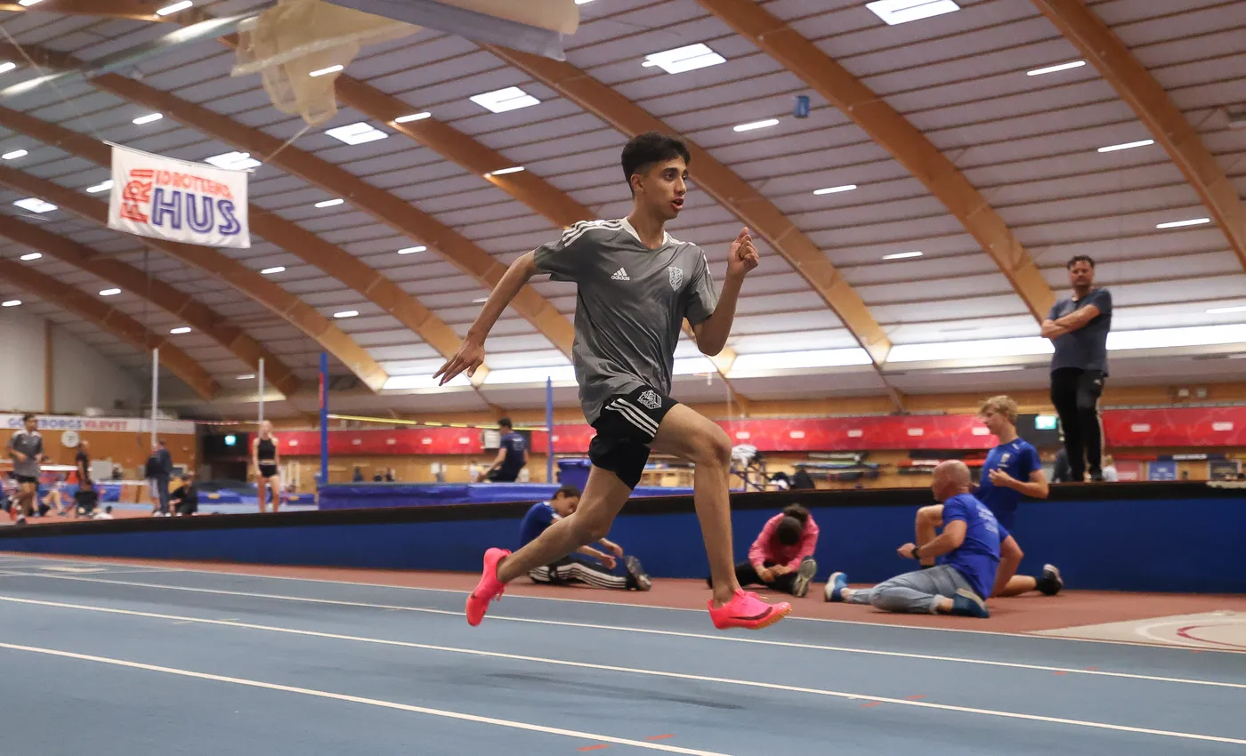 شخص شاب يرتدي زي رياضي رمادي مع شورت أسود يجري بسرعة على مضمار داخلي. يظهر في الخلفية عدد من الرياضيين الآخرين، بينما توجد لافتة معلقة تشير إلى اسم المكان.