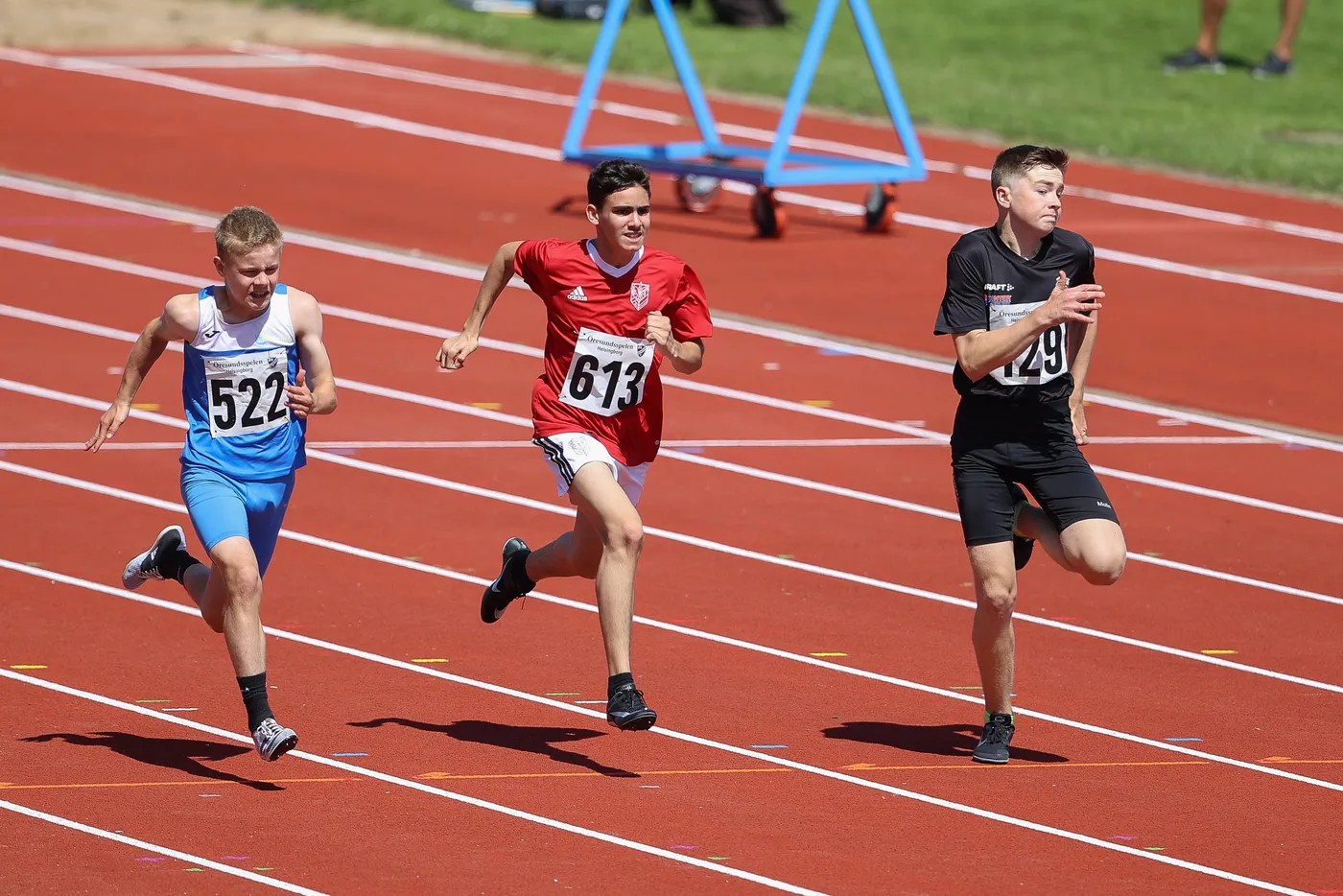 ثلاثة شبان يتنافسون في سباق جري على المضمار الأحمر، يرتدي أحدهم قميصاً أزرق برقم 522، بينما الآخر يرتدي قميصاً أحمر يحمل الرقم 613، والشاب الثالث يرتدي ملابس سوداء برقم 129. يظهرون في حركة سريعة أثناء السباق.