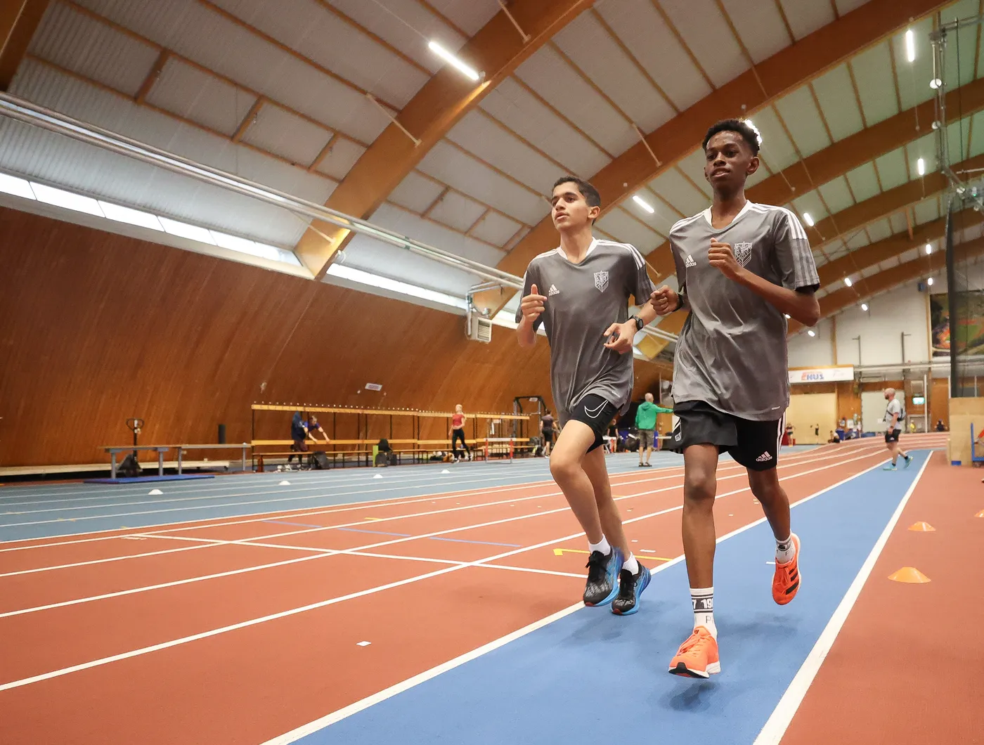 شبان يرتديان قمصان رياضية رمادية قصيرة الأكمام، يقومان بالجري على مضمار في قاعة مغلقة، بينما يظهر أشخاص آخرون يمارسون أنشطة رياضية في الخلفية. تُعطي الصور شعوراً بالحيوية والنشاط.