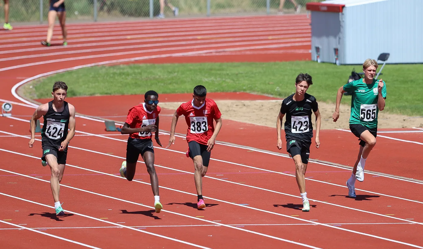 خمسة عدائين يرتدون زياً رياضياً مختلفاً يتنافسون في سباق على مضمار رياضي. يتميز المضمار بلونه الأحمر، وتظهر خلفهم حلبة سباق وأشخاص آخرون يشاهدون.