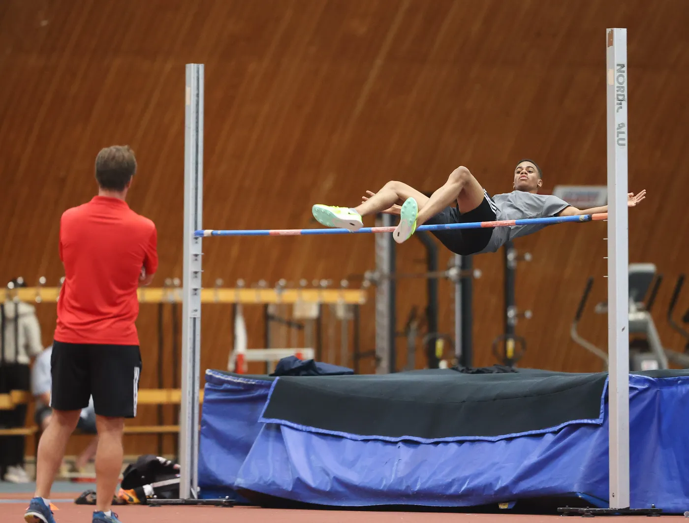 يظهر ممارس رياضي خلال قفز عالي، حيث يتمتع بوضعية مرتفعة فوق العارضة. يبدو المدرب في الخلف يتابع التركيز على الأداء.