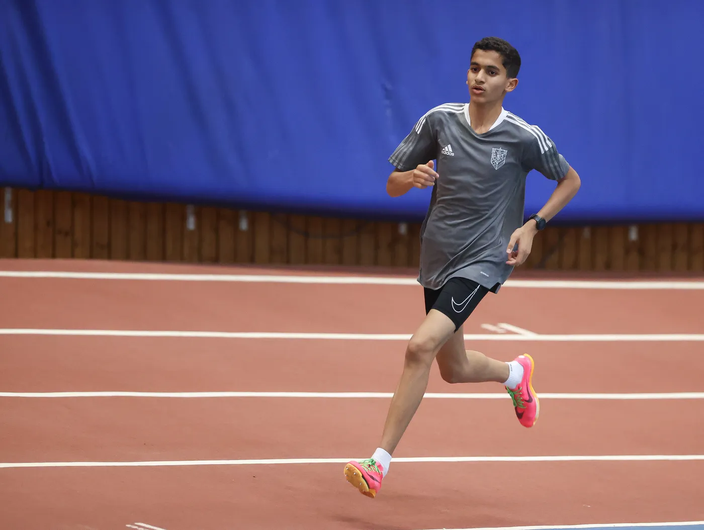 شخص شاب يركض بجدية على مضمار داخلي، وهو يرتدي تيشيرت رمادي وشورت قصير، وحذاء رياضي بألوان زاهية. خلفه جدار أزرق، ويظهر التركيز على وجهه.