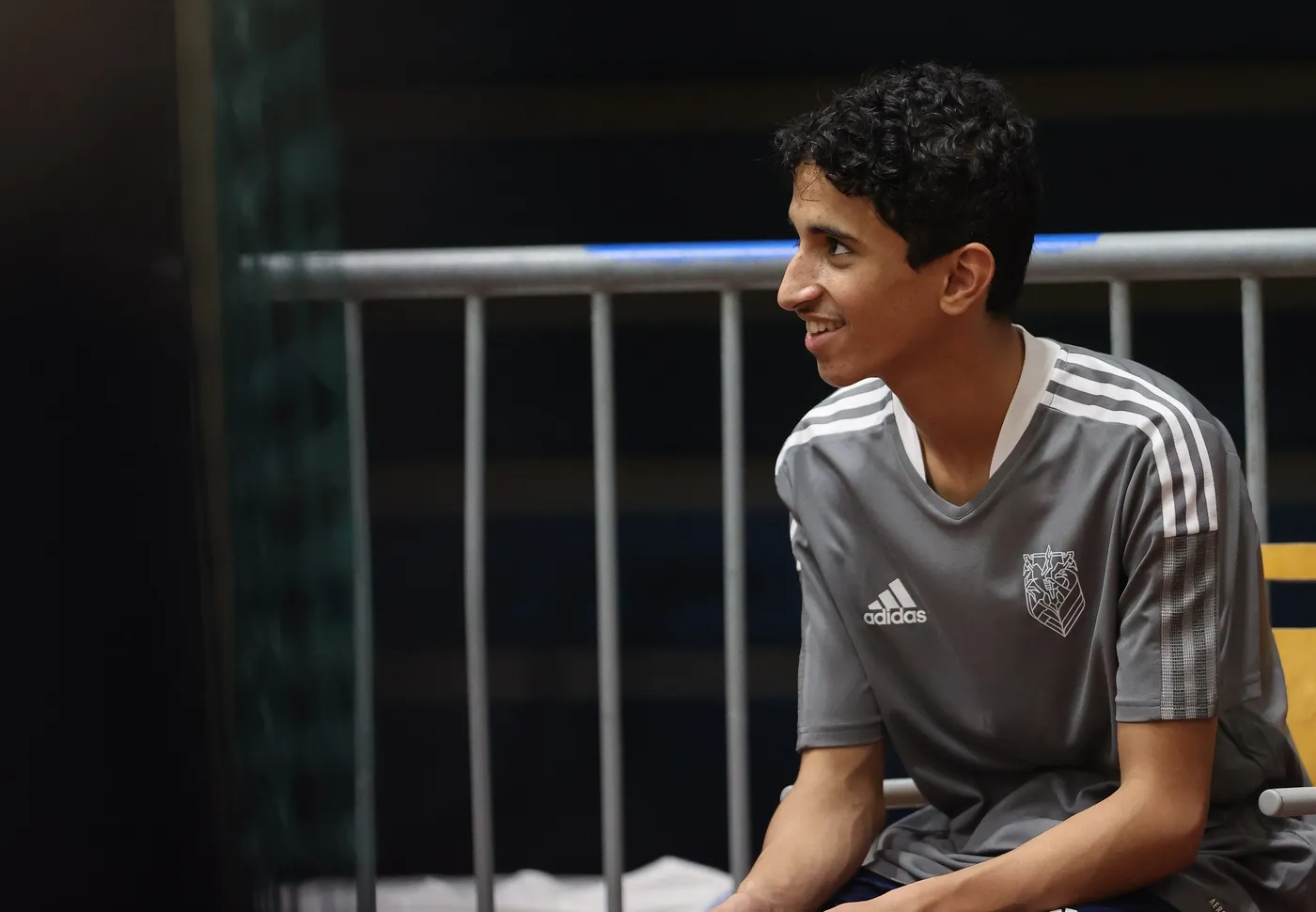 شاب يجلس على مقعد، مبتسمًا ويبدو عليه الانتباه. يرتدي قميص رياضي رمادي يحمل شعار فريق، والخلفية تحتوي على سياج معدني وأجواء رياضية.