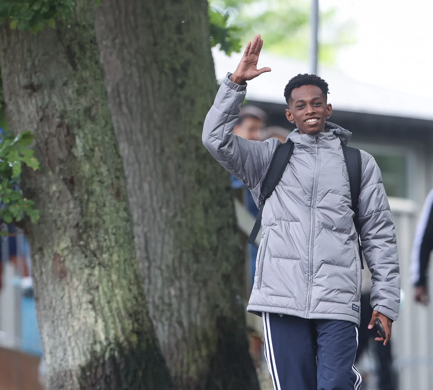 شاب يرتدي معطفًا رماديًا ويستعرض ابتسامة عريضة أثناء تحيته بيده، ويظهر في الخلفية شجرة كبيرة وبعض الأشخاص. يبدو أنه في طريقه لمكان ما.