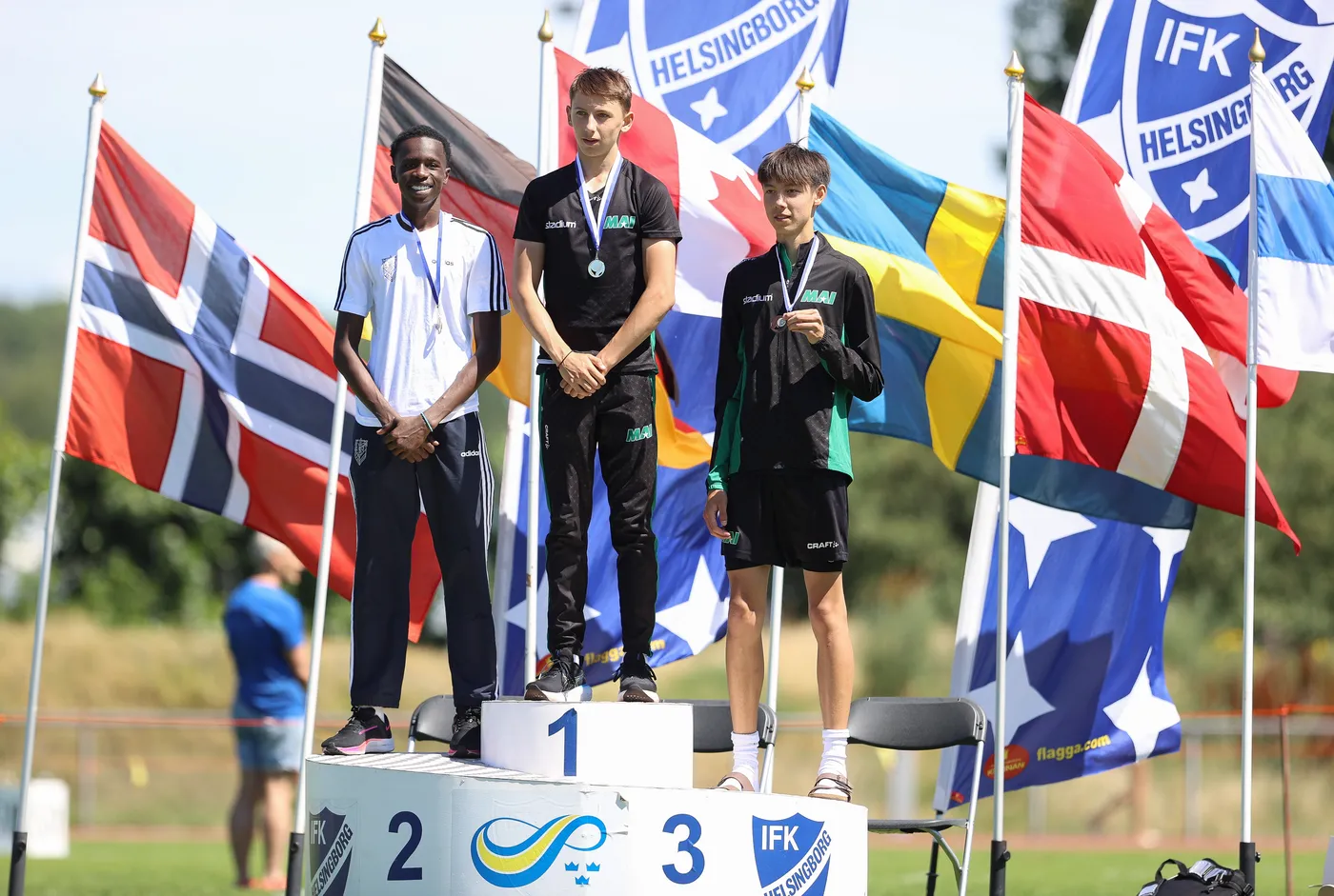 يظهر ثلاثة من لاعبي الرياضة يقفون على منصة التتويج، حيث يحمل الأول ميدالية ذهبية والثاني فضية والثالث برونزية. في الخلفية، تلوح أعلام عدة دول، مما يشير إلى حدث دولي.