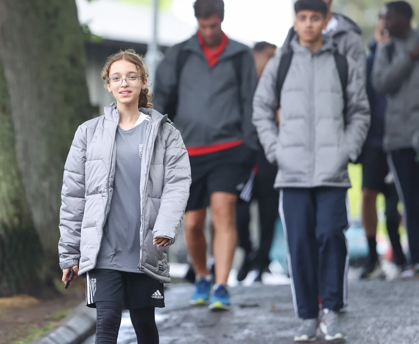 فتاة ترتدي سترة رمادية وسروال قصير أسود تسير مبتسمة في طريق لحديقة أو ملعب، بينما خلفها مجموعة من الشباب يرتدون ملابس رياضية متشابهة. الأشجار تحيط بالمكان مما يعطي شعورًا بالهدوء.