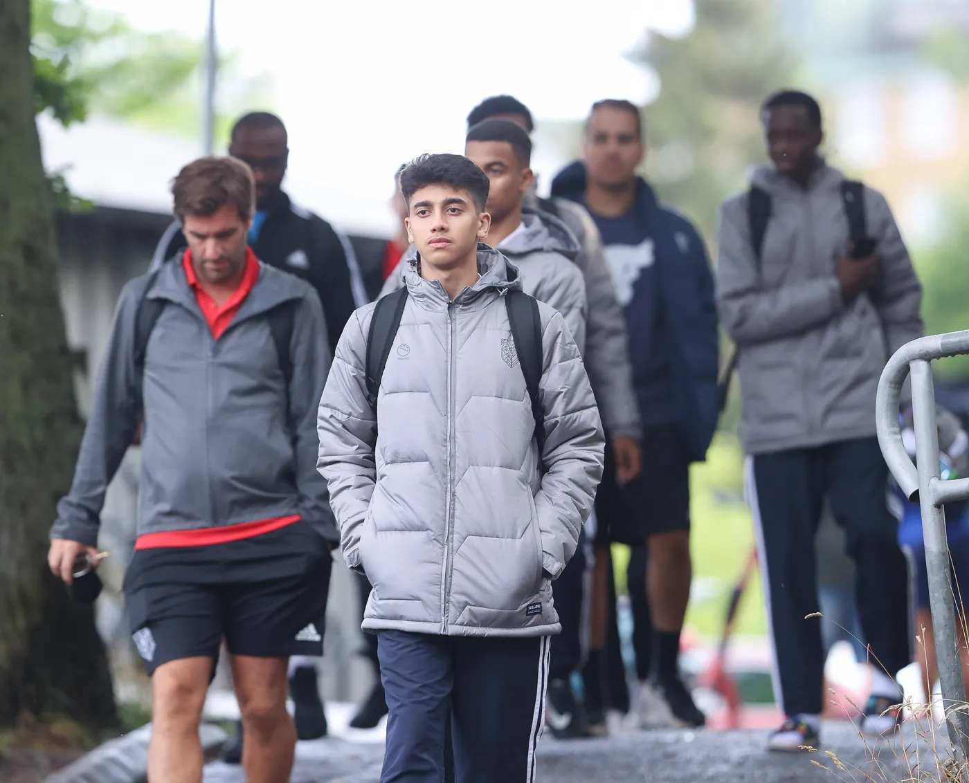 مجموعة من الشبان يرتدون سترات رمادية وملابس رياضية تسير على الرصيف، حيث يبدو أنهم متجهون إلى وجهة معينة. أحدهم في المقدمة يبدو جادًا بينما الآخرون يتواجدون خلفه.