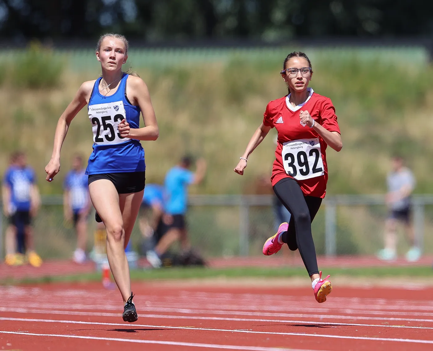 متسابقات في سباق عداء على المضمار، ترتدي إحداهن زيًا أزرق برقم 25 والأخرى زيًا أحمر برقم 392. يظهر الحماس والتنافس بينهما في يوم مشمس.